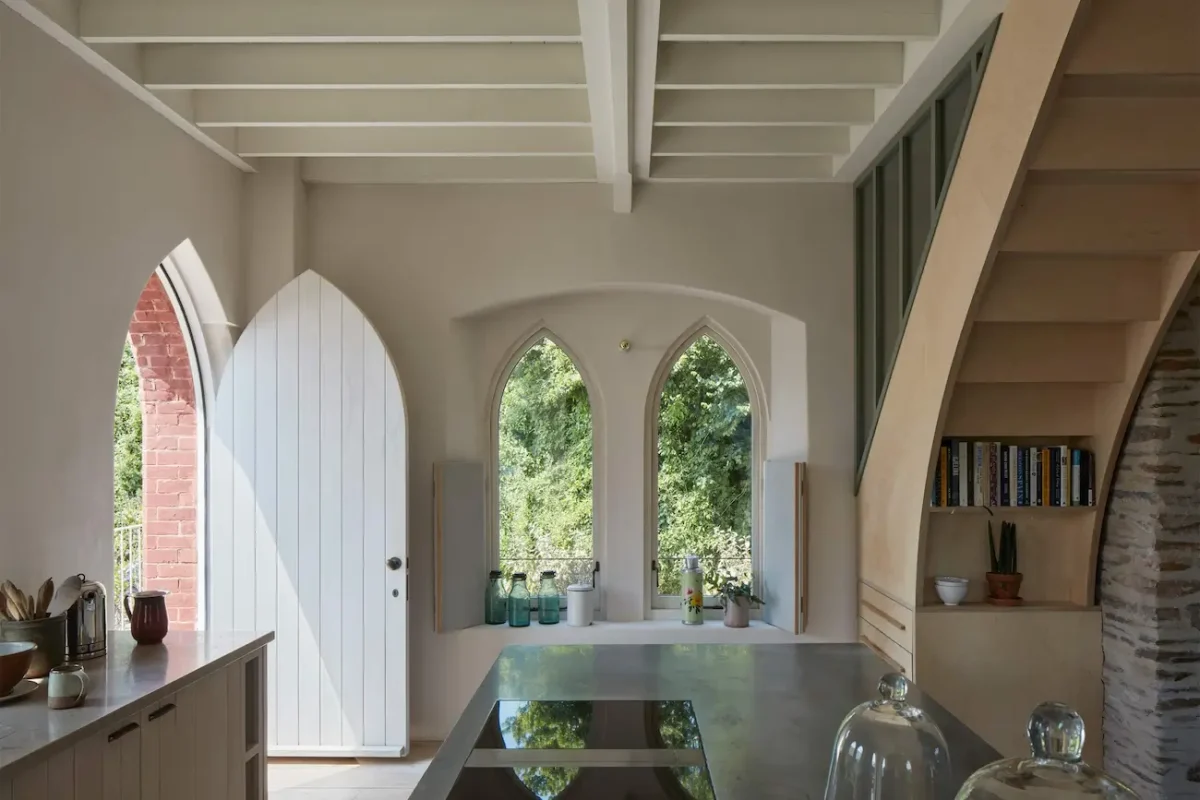 Kitchen inside The Old Chapel by Tuckey Design Studio featuring Gothic-style arched windows, a polished zinc countertop, and a plywood staircase leading to a mezzanine level.