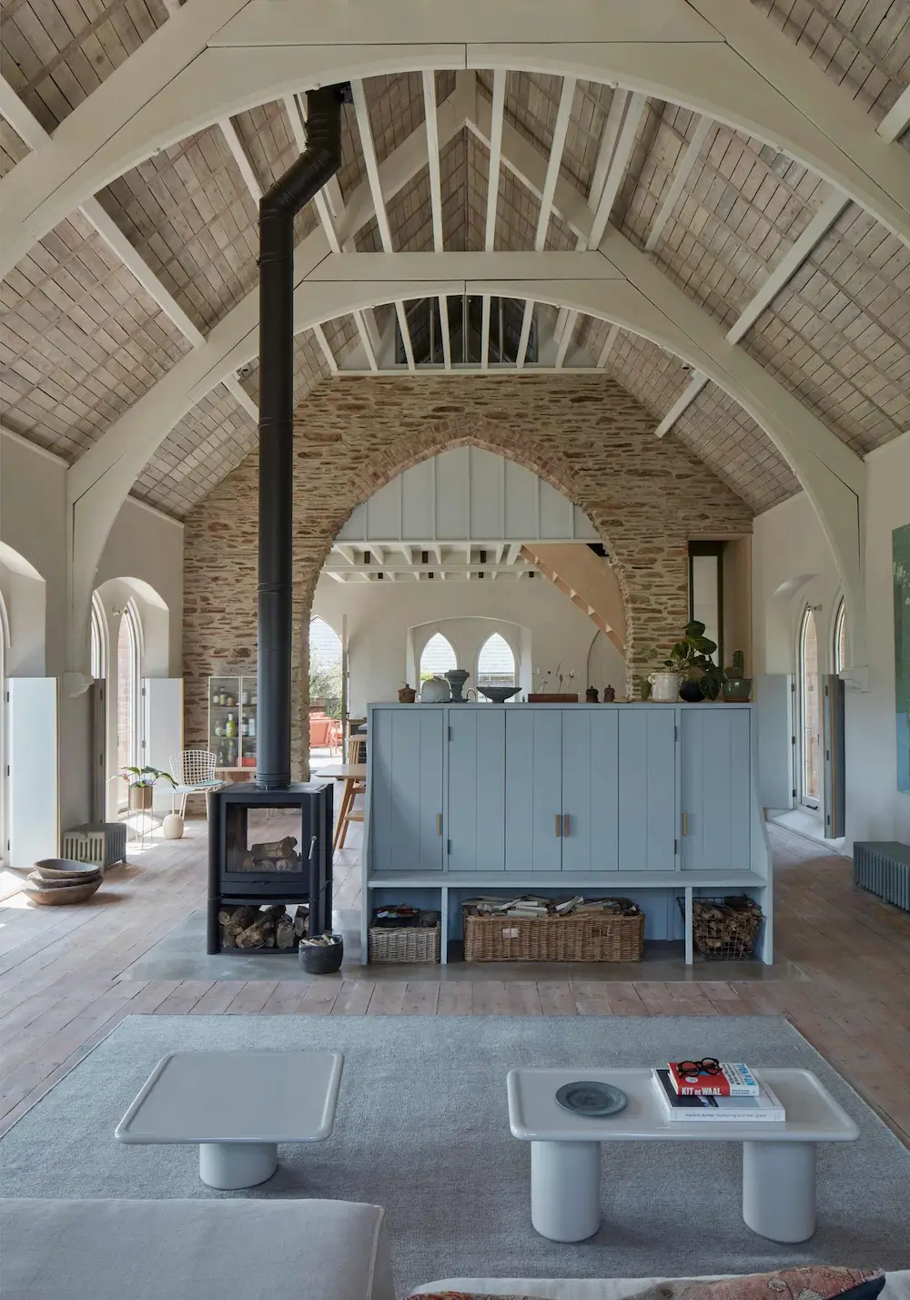 Open-plan living area inside a converted Devon chapel with exposed stone arches, vaulted timber ceilings, and a central wood-burning stove.