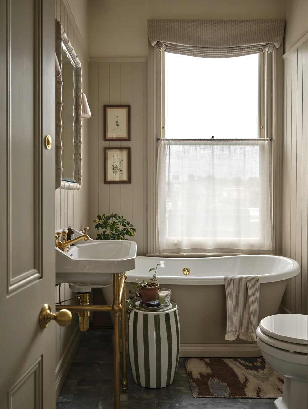 Traditional Victorian bathroom with a freestanding bathtub beneath a window, a wall-mounted sink with brass fittings, vertical paneling, and soft neutral finishes.