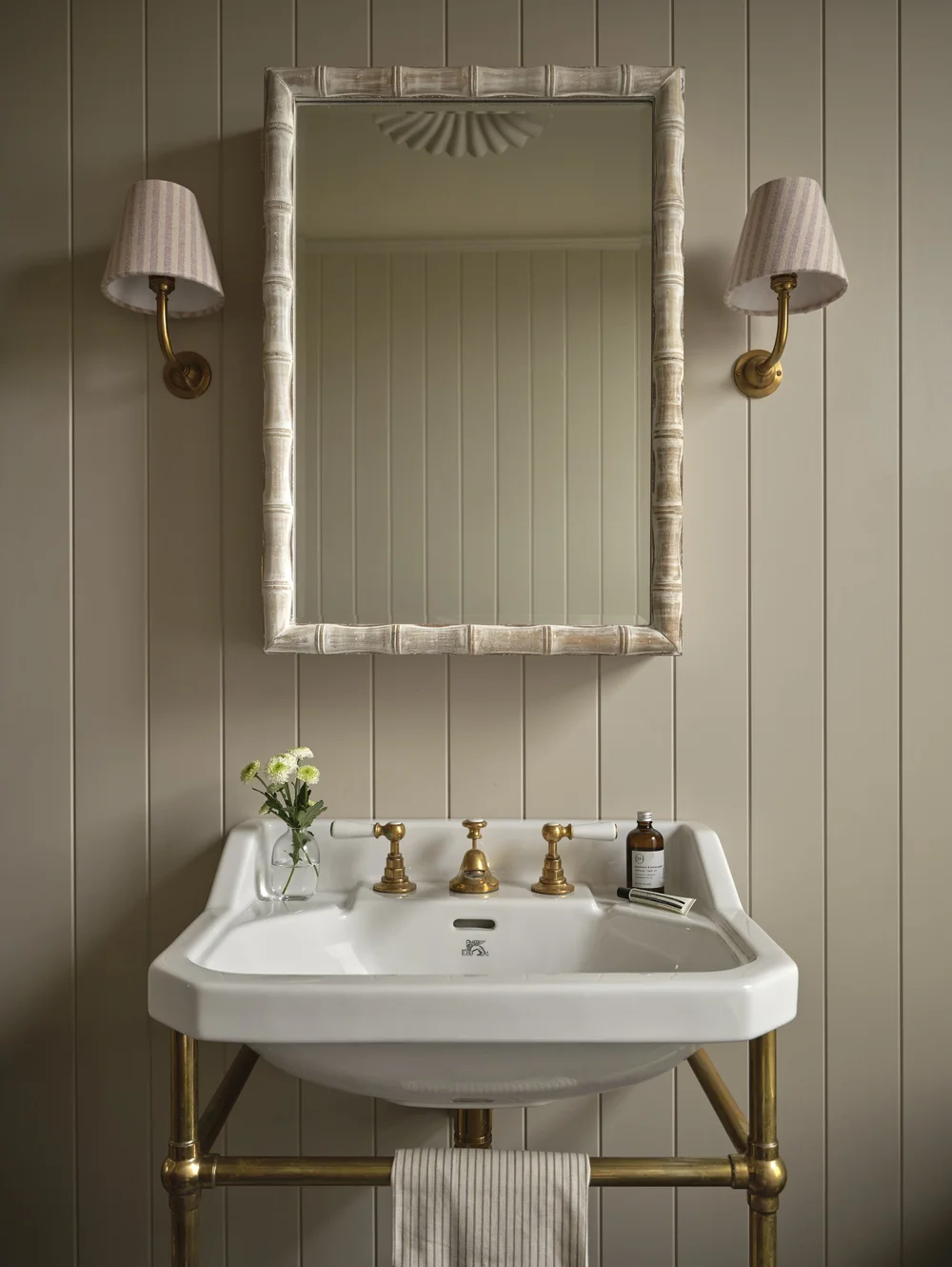 Traditional wall-mounted bathroom sink with exposed brass plumbing, tongue-and-groove paneling, a bamboo-style mirror, and brass sconces with pleated fabric shades.