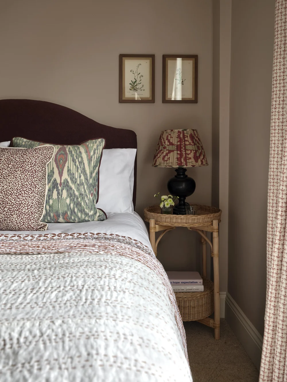 Cozy bedroom corner with a burgundy upholstered headboard, patterned accent pillows, a woven rattan bedside table, a pleated table lamp, and botanical artwork.