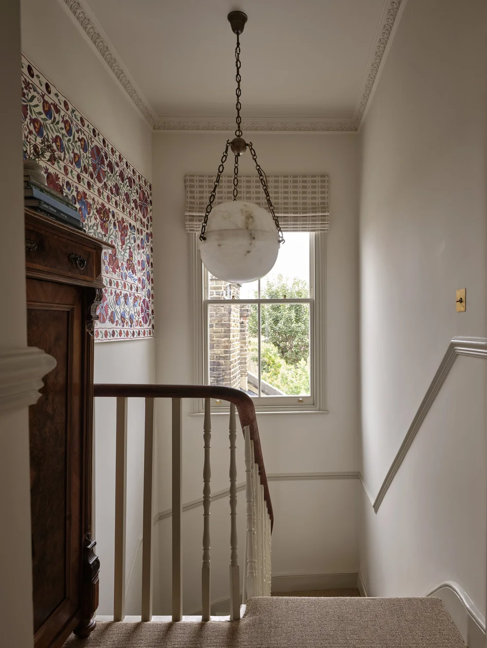 Victorian staircase landing with original balustrade, ornate ceiling cornices, a globe pendant light, a patterned textile wall hanging, and a large sash window.