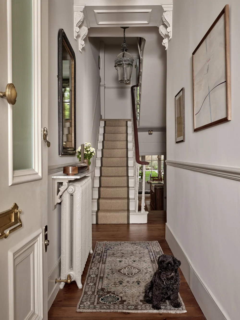 Victorian townhouse hallway in Hackney featuring original decorative archway, ornate corbels, a cast-iron radiator, staircase with a neutral runner, vintage rug, and a black dog resting in the entry.