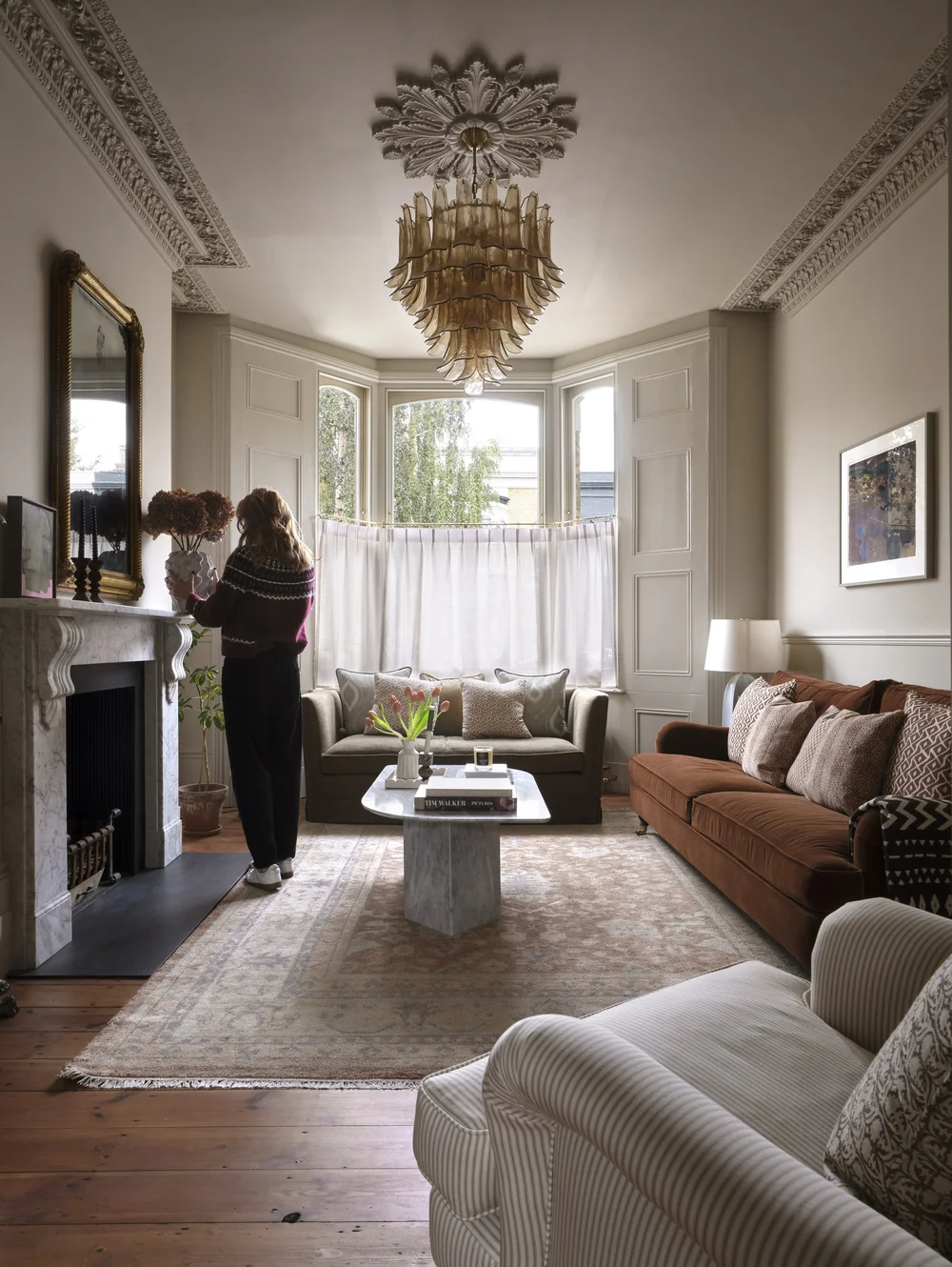 Elegant Victorian living room by Frank and Faber with ornate ceiling moldings, a marble fireplace, a Murano-style chandelier, rust-colored velvet sofa, bay window, and vintage rug.