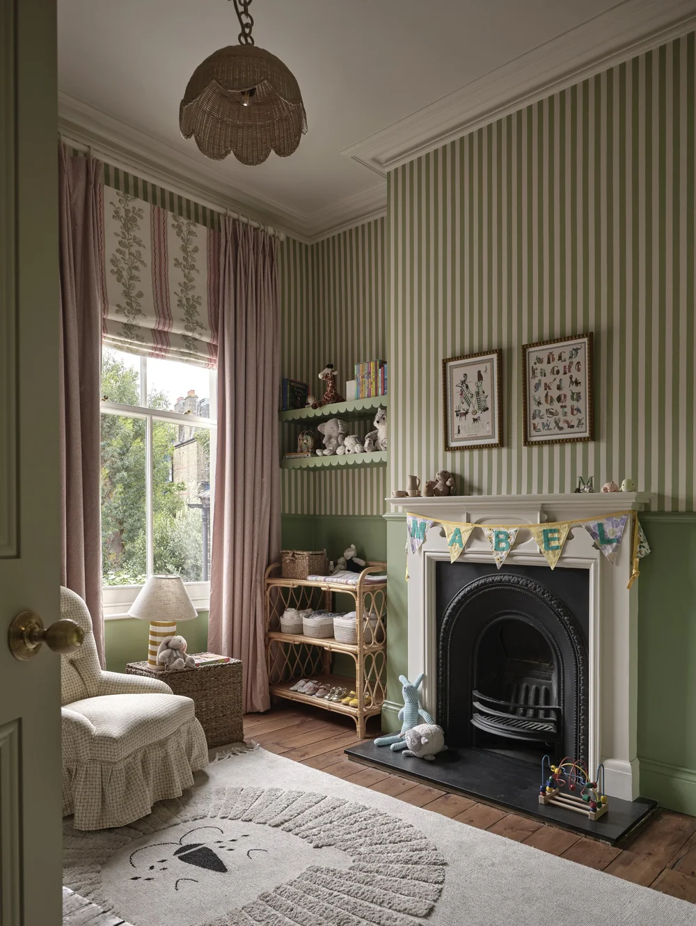 Victorian nursery with green striped wallpaper, original fireplace, blush pink curtains, a rattan toy shelf, vintage-style decor, and a playful lion rug.