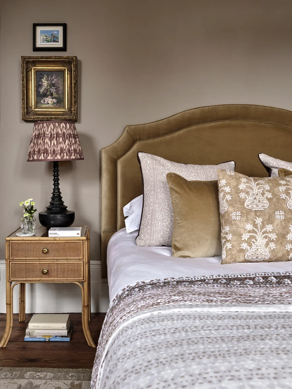 Warm neutral bedroom with an olive velvet upholstered headboard, layered patterned pillows, a rattan nightstand, a pleated table lamp, and vintage artwork.