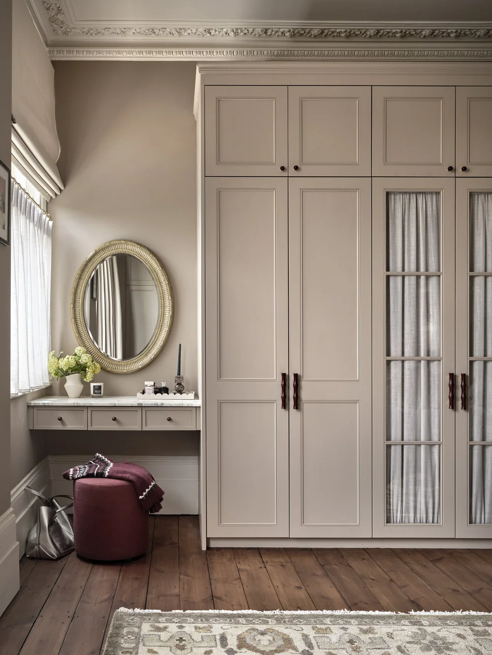 Custom built-in wardrobes and dressing area in a Victorian bedroom featuring warm greige shaker cabinetry, a marble-topped vanity, an oval mirror, and original ornate ceiling cornices.