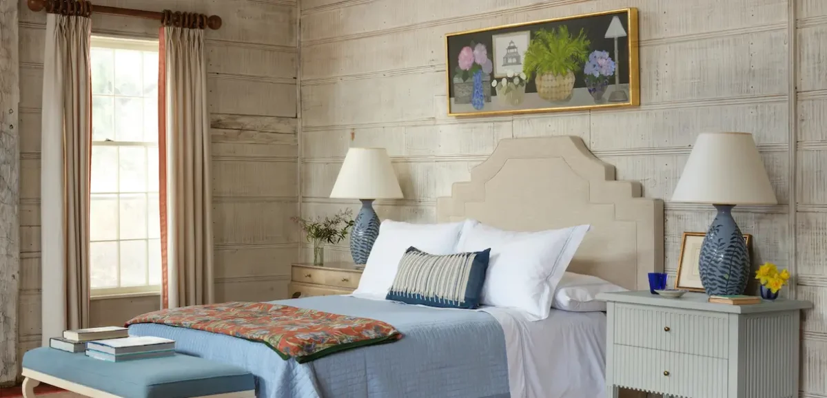 Bright bedroom designed in Bunny Williams' layered style, featuring rustic wood plank walls, a geometric upholstered headboard, blue ceramic lamps, crisp white bedding, and a mix of traditional and contemporary furnishings.