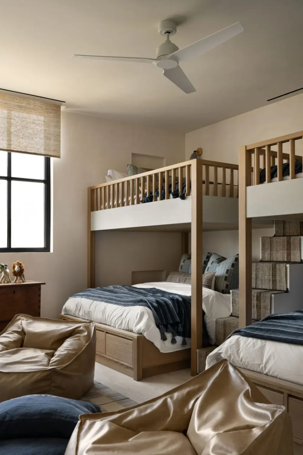 Bright bunk room featuring two custom wood-and-white bunk beds with integrated storage drawers and carpeted stairs, styled with blue plaid bedding, oversized bean bag chairs, a large black-framed window, and warm neutral finishes for a relaxed family retreat.