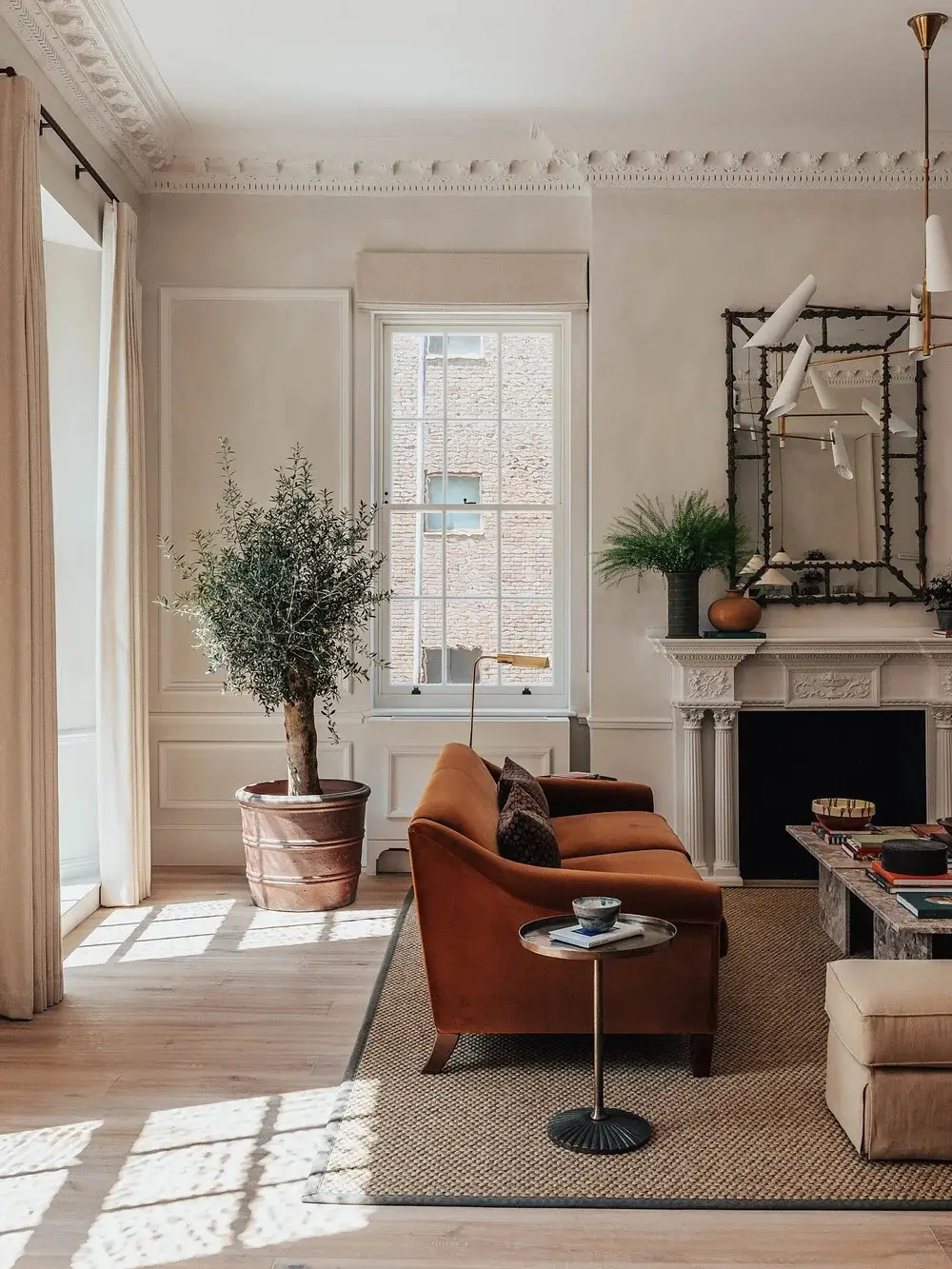 Bright sitting area with rust velvet sofa, olive tree, ornate fireplace, and tall sash windows in a luxury Mayfair apartment.