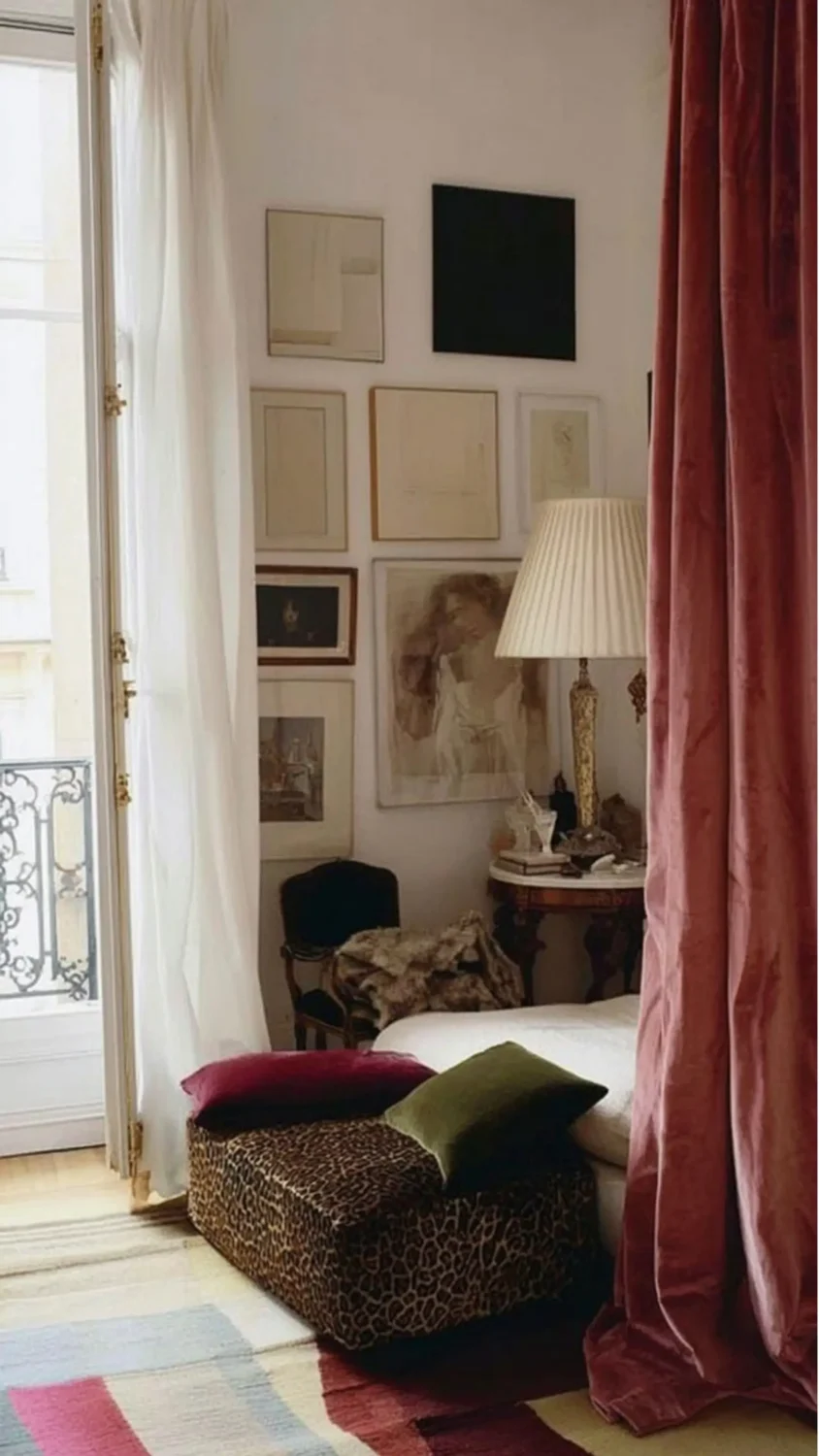 Elegant Parisian bedroom with floor-to-ceiling dusty rose velvet curtains, a curated gallery wall of vintage artwork, a leopard-print bench with jewel-toned velvet cushions, and soft natural light streaming through French doors.