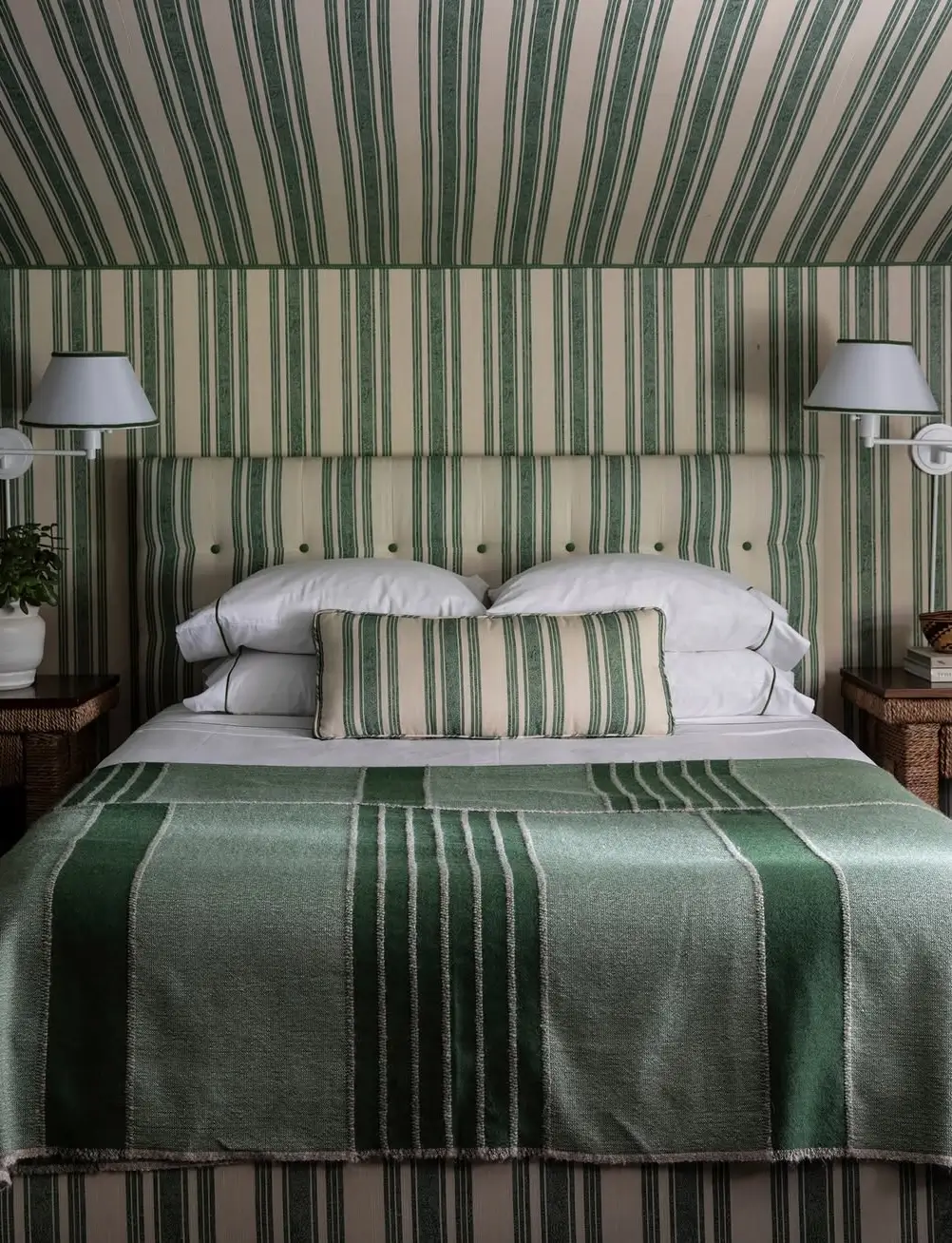 Cozy guest bedroom by Mark D. Sikes featuring green-and-white striped wallpaper, a matching upholstered headboard, and layered striped textiles.