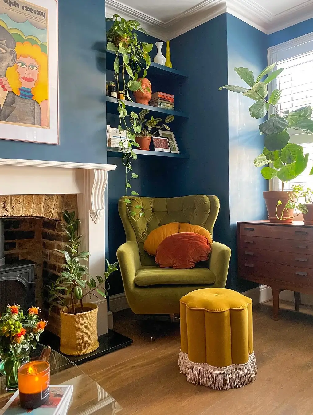 Eclectic living room in Ola Zwolenik's home featuring deep blue walls, a green velvet wingback chair, colorful cushions, abundant houseplants, and a mustard fringed ottoman.