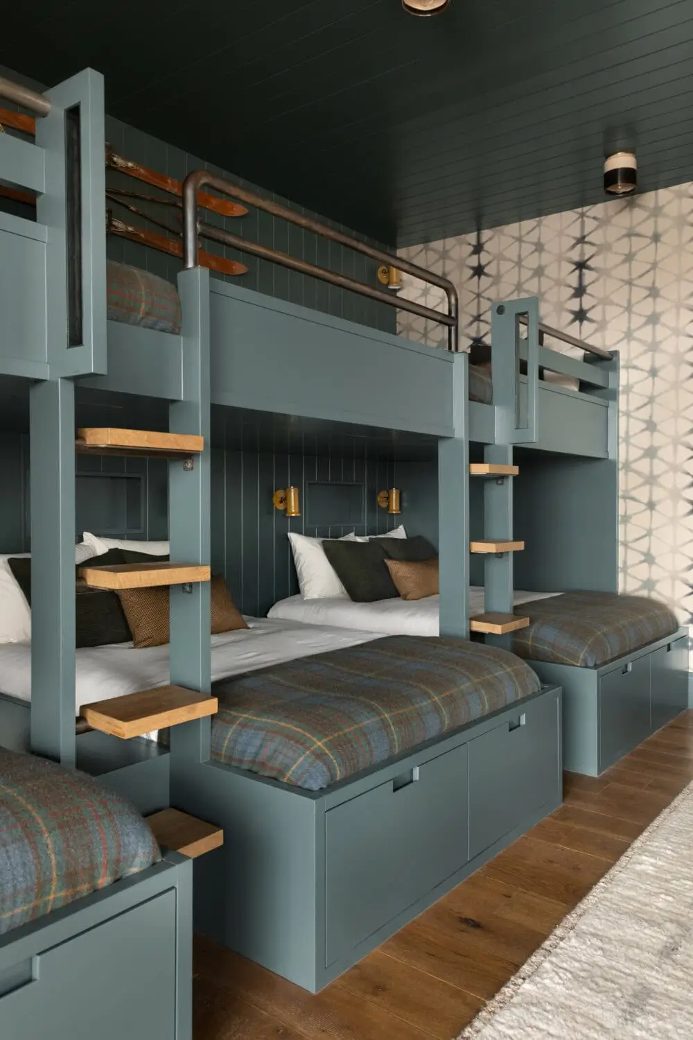Built-in bunk room by Studio McGee featuring four custom blue-gray bunk beds with integrated storage drawers, wood ladder treads, and metal safety rails. The moody space includes dark paneled walls and ceiling, plaid bedding, warm brass reading lights, patterned wallpaper, and wide-plank wood flooring, creating a cozy, cabin-inspired retreat.