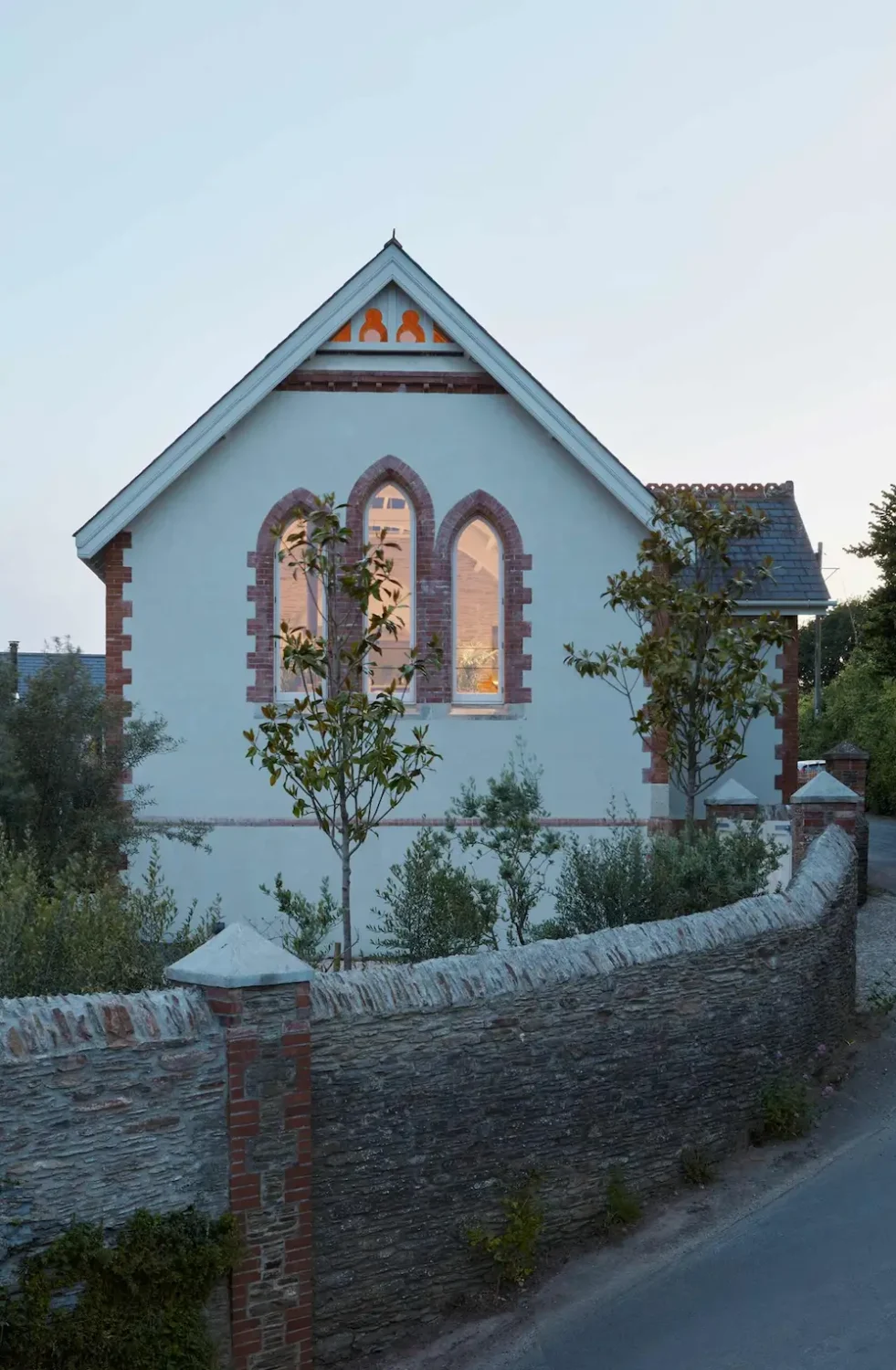Exterior view of The Old Chapel by Tuckey Design Studio in west Devon, featuring Gothic-style arched windows, white rendered walls, red brick detailing, and a traditional stone boundary wall at dusk.