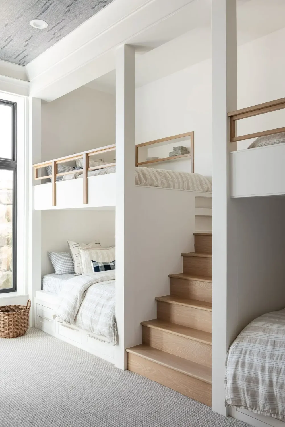 Bright custom bunk room featuring four built-in white bunk beds arranged around a central wood staircase, with integrated storage drawers, light plaid bedding, wood-accent railings, and a large black-framed window. The airy space combines crisp white finishes, soft textures, and warm wood details for a clean, modern retreat.