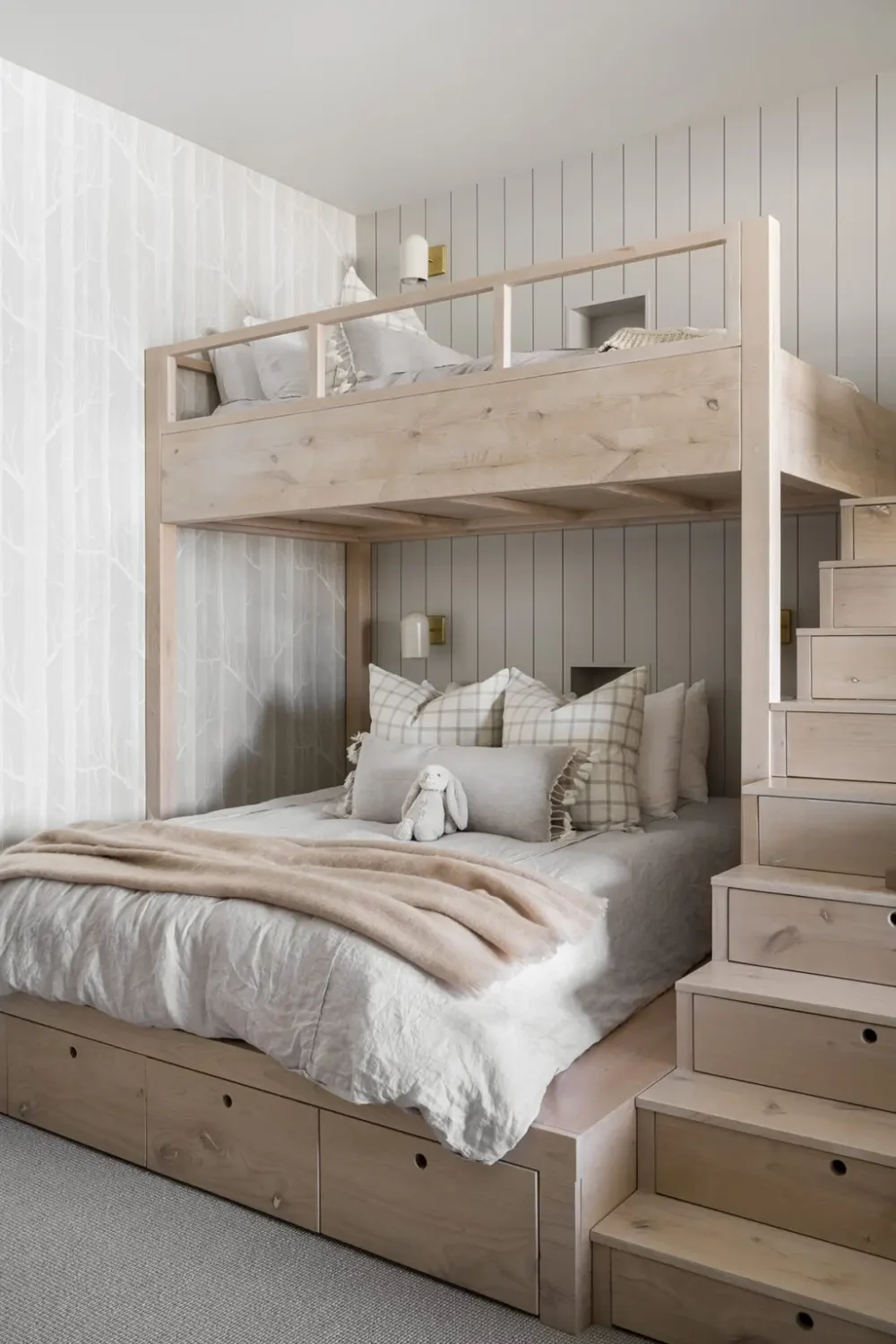 Custom light wood bunk bed with a queen-size lower bed and twin upper bunk, featuring built-in storage drawers, staircase access, paneled walls, subtle tree-pattern wallpaper, and soft neutral bedding in a serene, Scandinavian-inspired bedroom.
