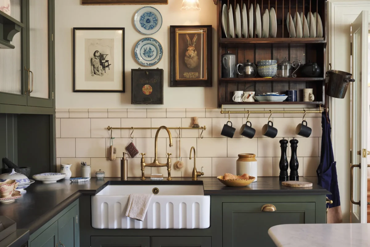 Victorian deVOL kitchen sink with a white Belfast sink, aged brass bridge faucet, olive green cabinetry, cream subway tile backsplash, open plate rack, vintage artwork, and hanging black mugs.
