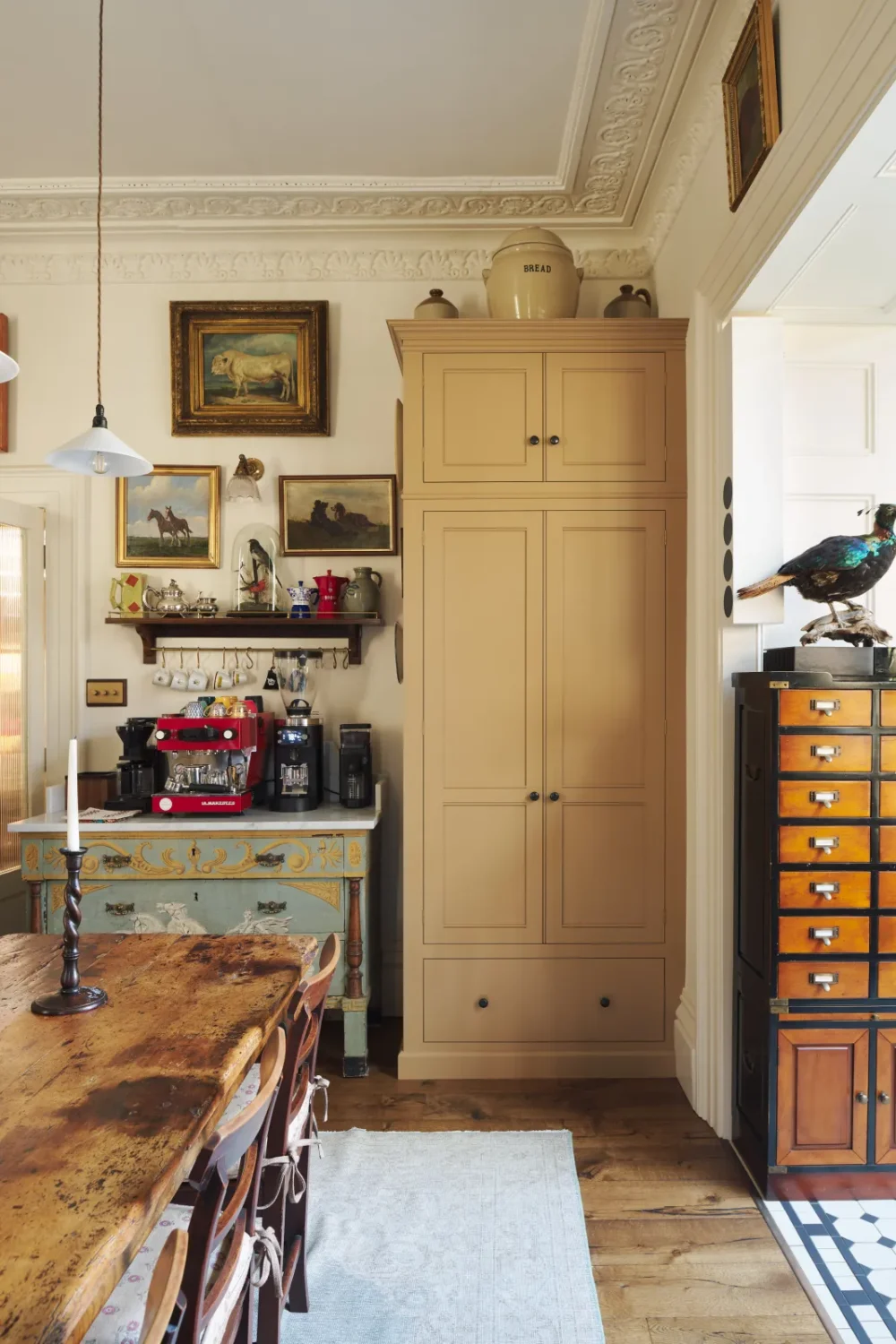 Eclectic Victorian kitchen dining area with a cream pantry cabinet, rustic farmhouse table, vintage coffee station, ornate plaster ceilings, antique artwork, and a restored apothecary cabinet topped with a taxidermy pheasant.
