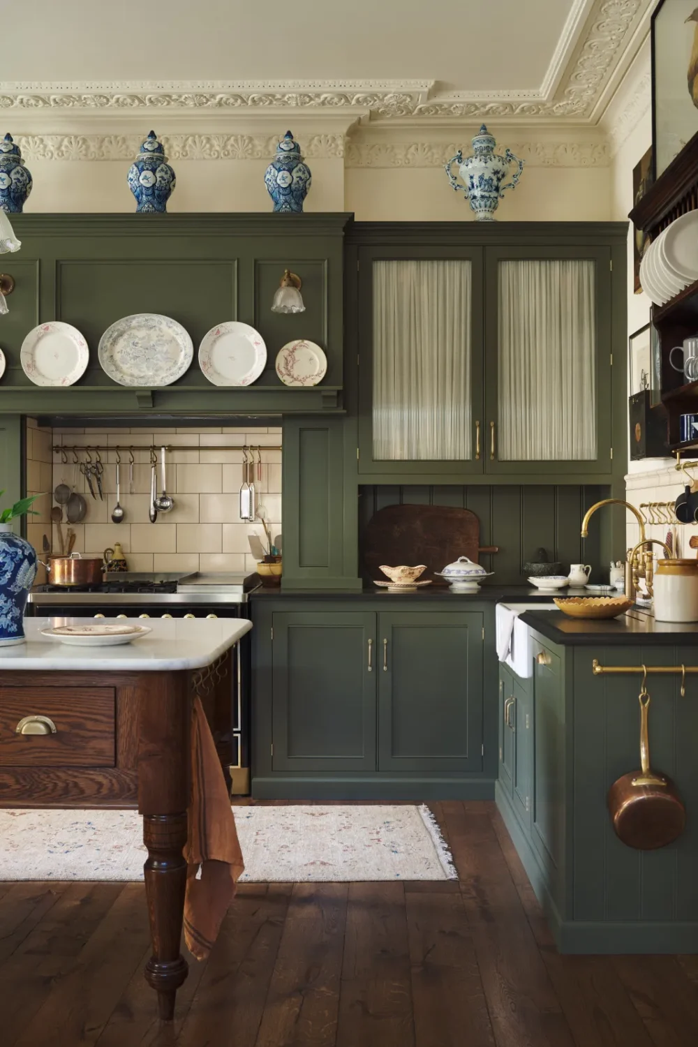 deVOL Victorian kitchen with olive green Shaker cabinets, a marble-topped oak island, blue-and-white porcelain display, brass fixtures, cream subway tiles, and ornate plaster ceilings.
