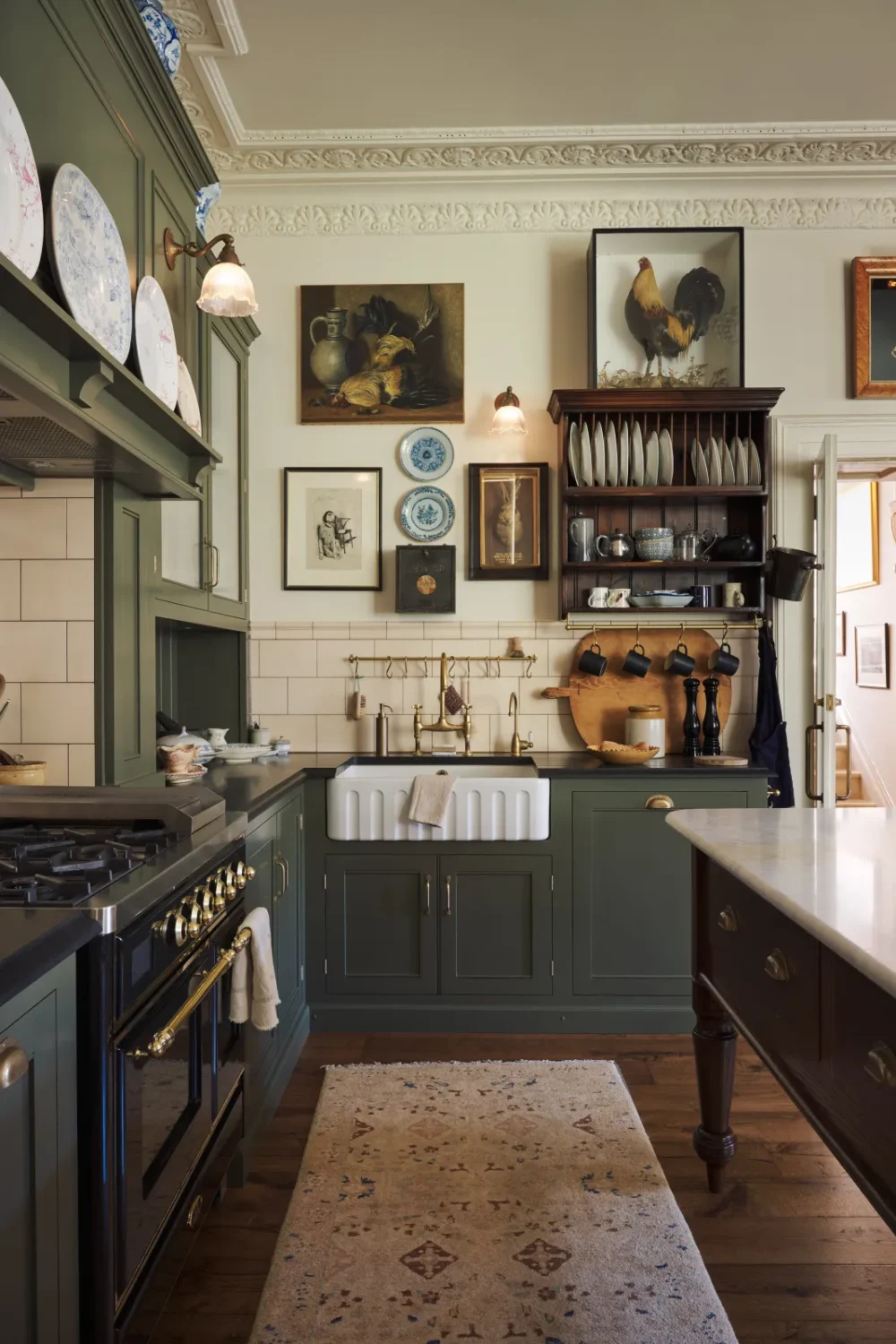 Eclectic deVOL Victorian kitchen with olive green cabinetry, a Belfast sink, brass bridge faucet, cream subway tiles, vintage artwork, an antique plate rack, and framed taxidermy.