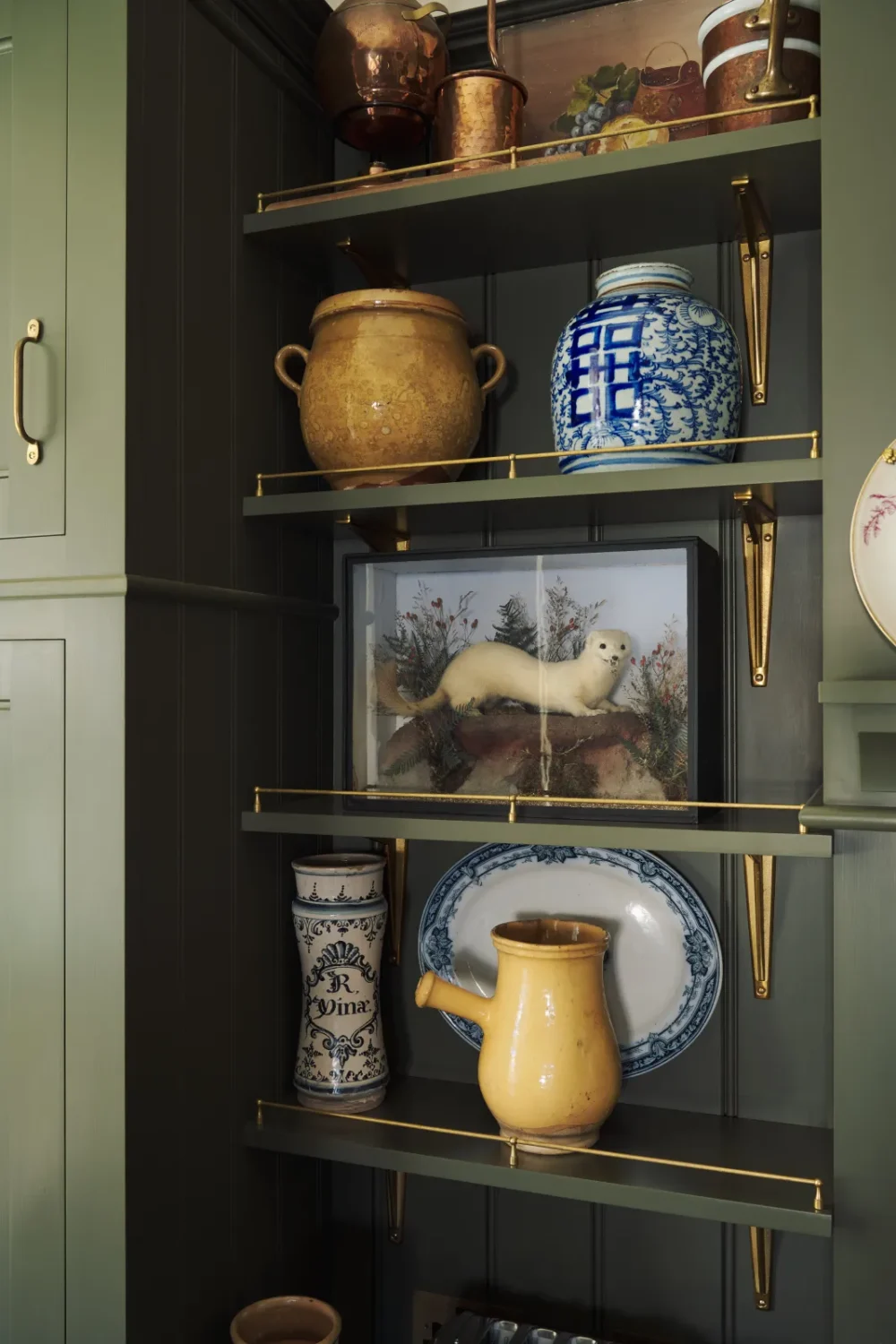 Open display shelves in a deVOL Victorian kitchen featuring blue-and-white porcelain, antique stoneware crocks, copper cookware, brass shelf rails, and a framed taxidermy display.