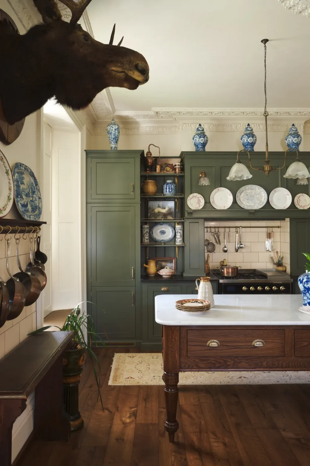 Eclectic Victorian kitchen by deVOL with olive green Shaker cabinets, a marble-topped oak island, blue-and-white porcelain, copper cookware, ornate plaster ceilings, and a mounted stag head.