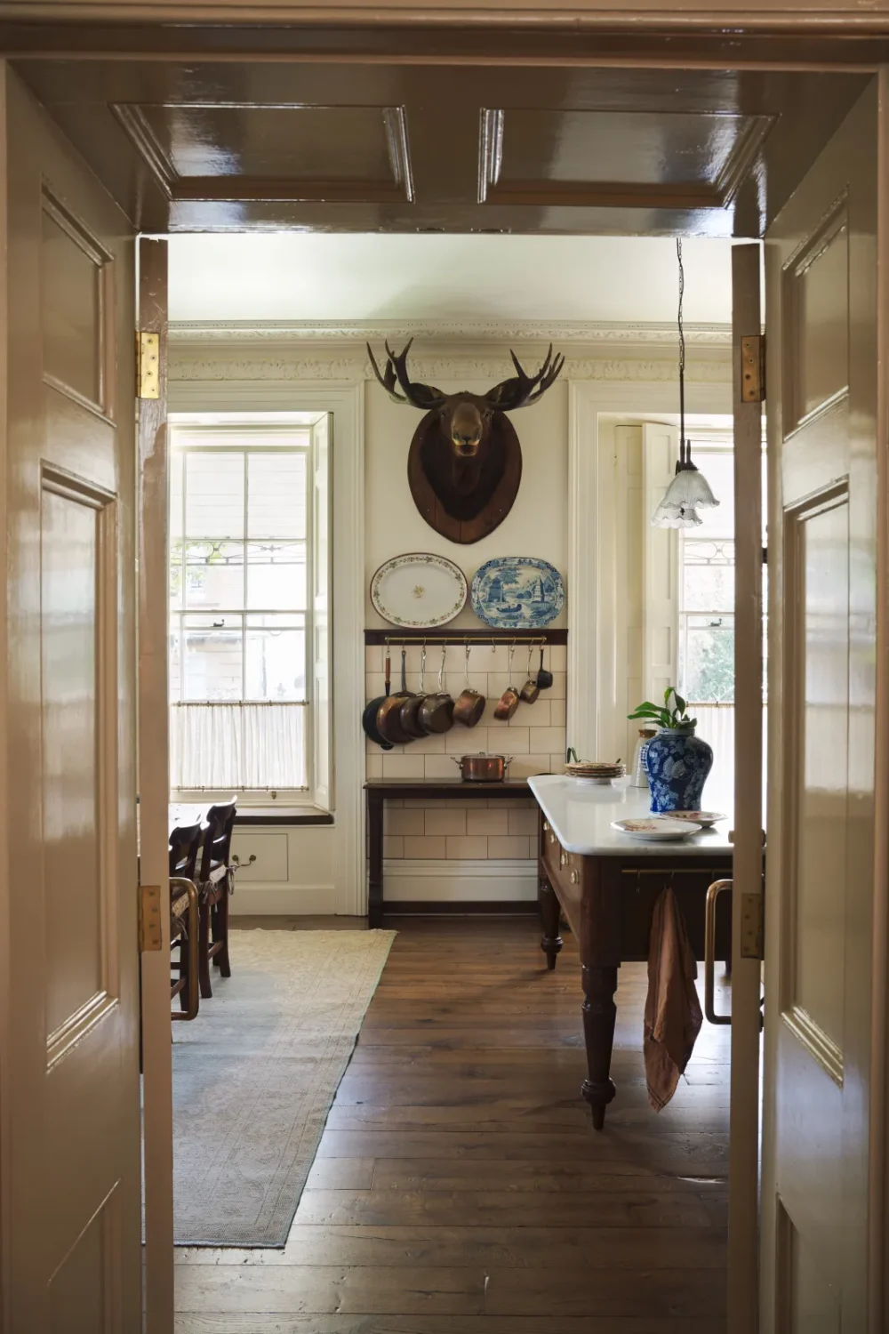 View through double doors into an eclectic deVOL Victorian kitchen featuring a marble-topped island, copper cookware, blue-and-white ceramics, tall sash windows, and a mounted stag head.