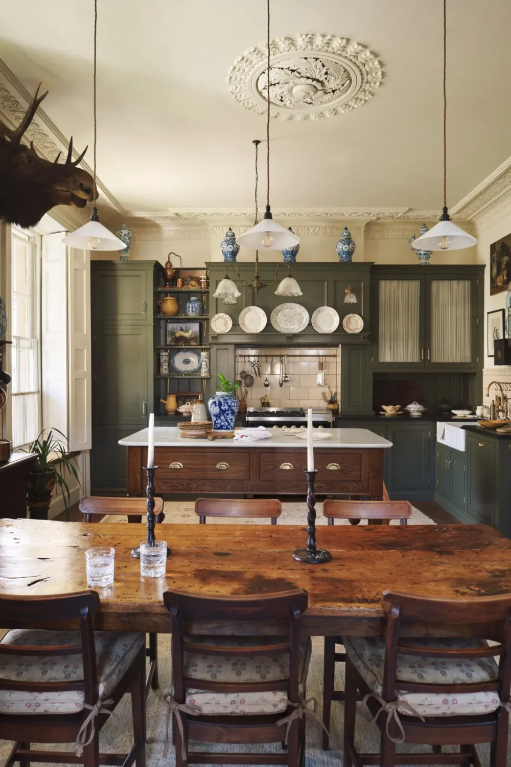 Eclectic Victorian kitchen by deVOL with olive green Shaker cabinetry, a marble-topped oak island, rustic farmhouse dining table, blue-and-white porcelain, ornate plaster ceiling, and a mounted stag head.