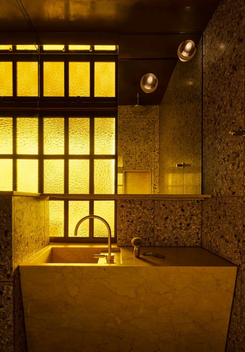 Moody bathroom in The Gentleman apartment by YSG Studio with terrazzo walls, stone vanity, textured Flemish glass window, and warm ambient lighting.