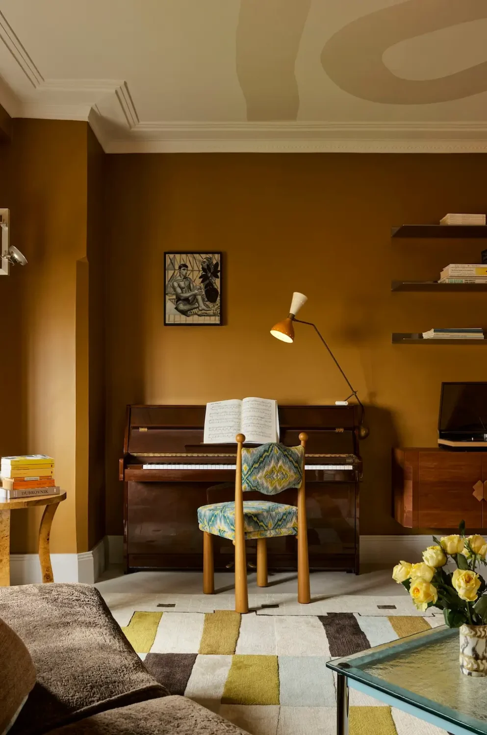 Music corner in The Gentleman apartment by YSG Studio with caramel walls, upright piano, patterned accent chair, floating shelves, and painted sculptural ceiling.