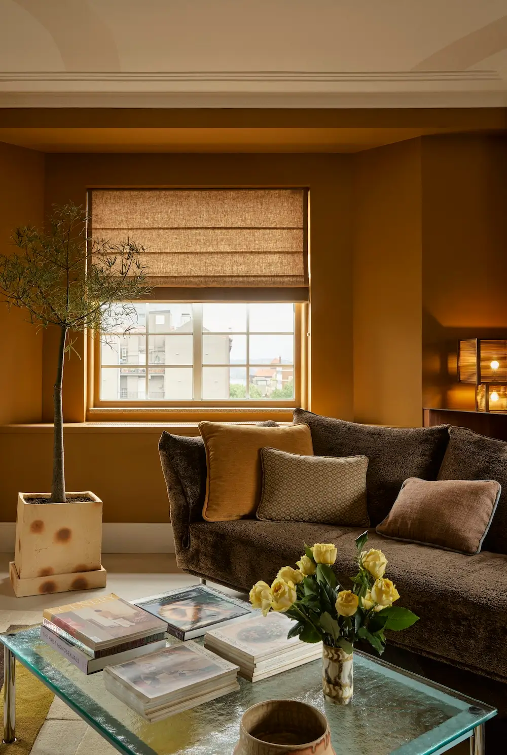 Warm living room in The Gentleman apartment by YSG Studio with caramel walls, deep brown sofa, linen Roman shade, olive tree, and glass coffee table.