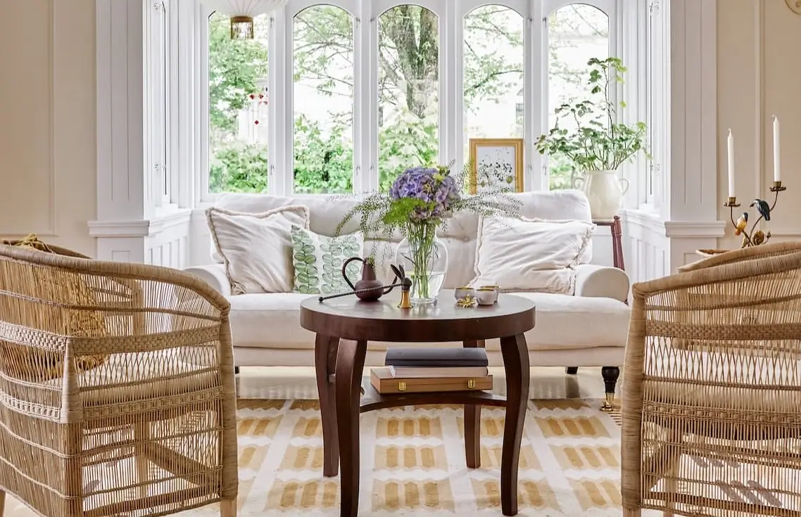 Bright traditional living room with two wicker lounge chairs, a cream linen sofa, a dark wood coffee table, and a patterned neutral rug in front of large bay windows.