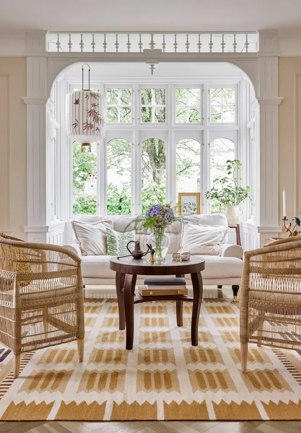 Traditional living room with wicker lounge chairs, a cream linen sofa, a dark wood coffee table, and a large bay window framed by decorative white millwork.