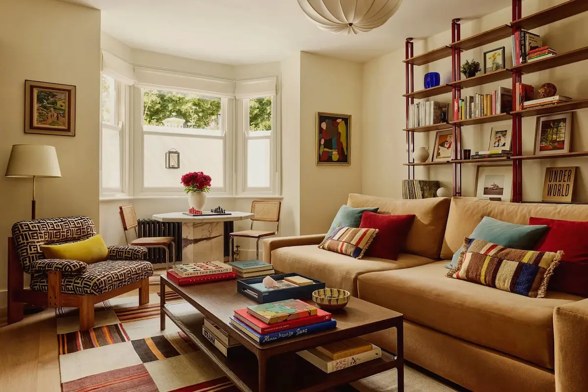 Warm living room in Michelle Dockery's London Victorian home featuring a neutral sofa with colorful cushions, a custom wall of open shelving, patterned armchair, striped rug, and a marble café table set within a bright bay window.