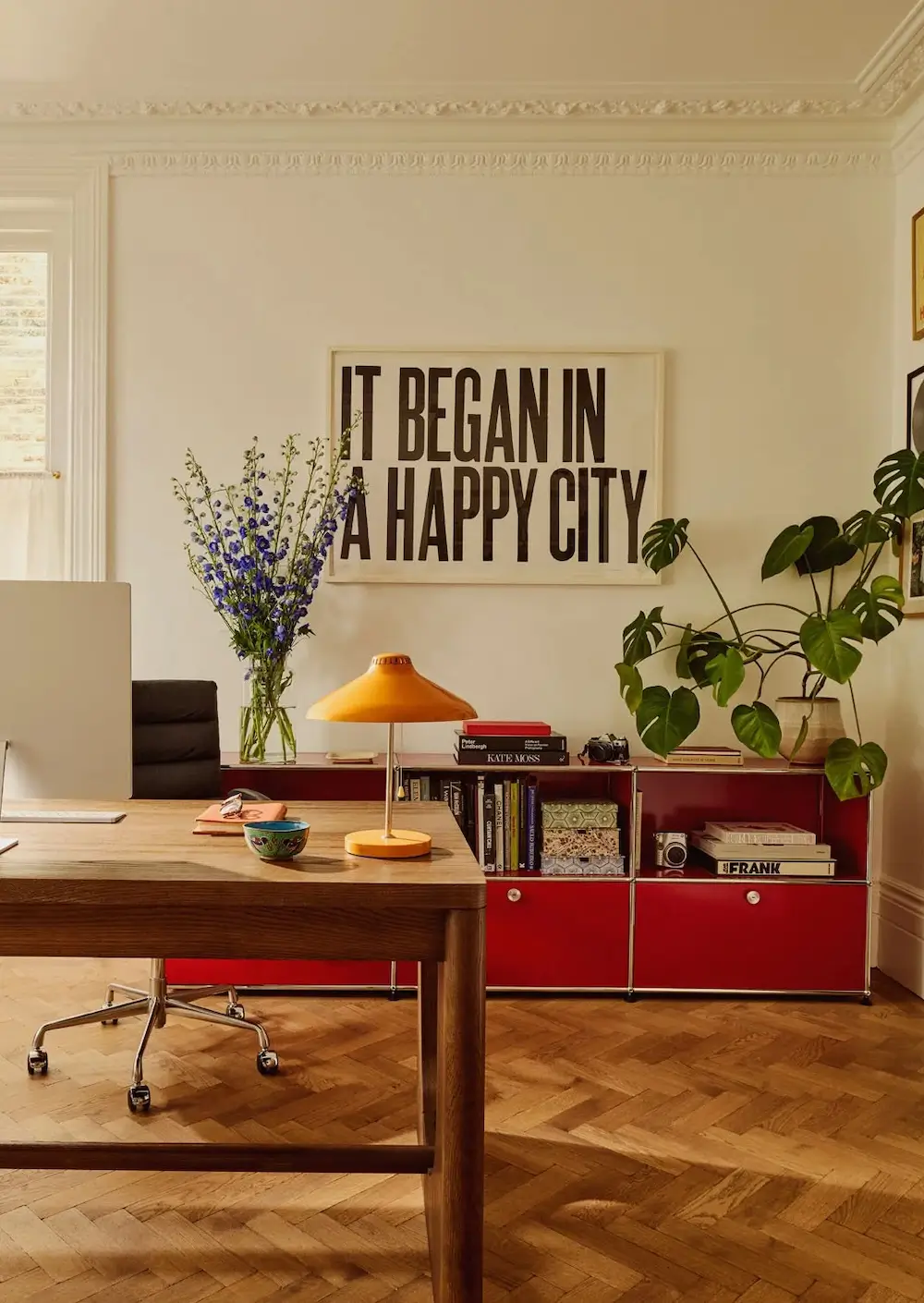 Home office in Michelle Dockery's London Victorian home featuring a walnut desk, red USM storage cabinet, herringbone wood floors, bold typography artwork, and lush indoor plants.