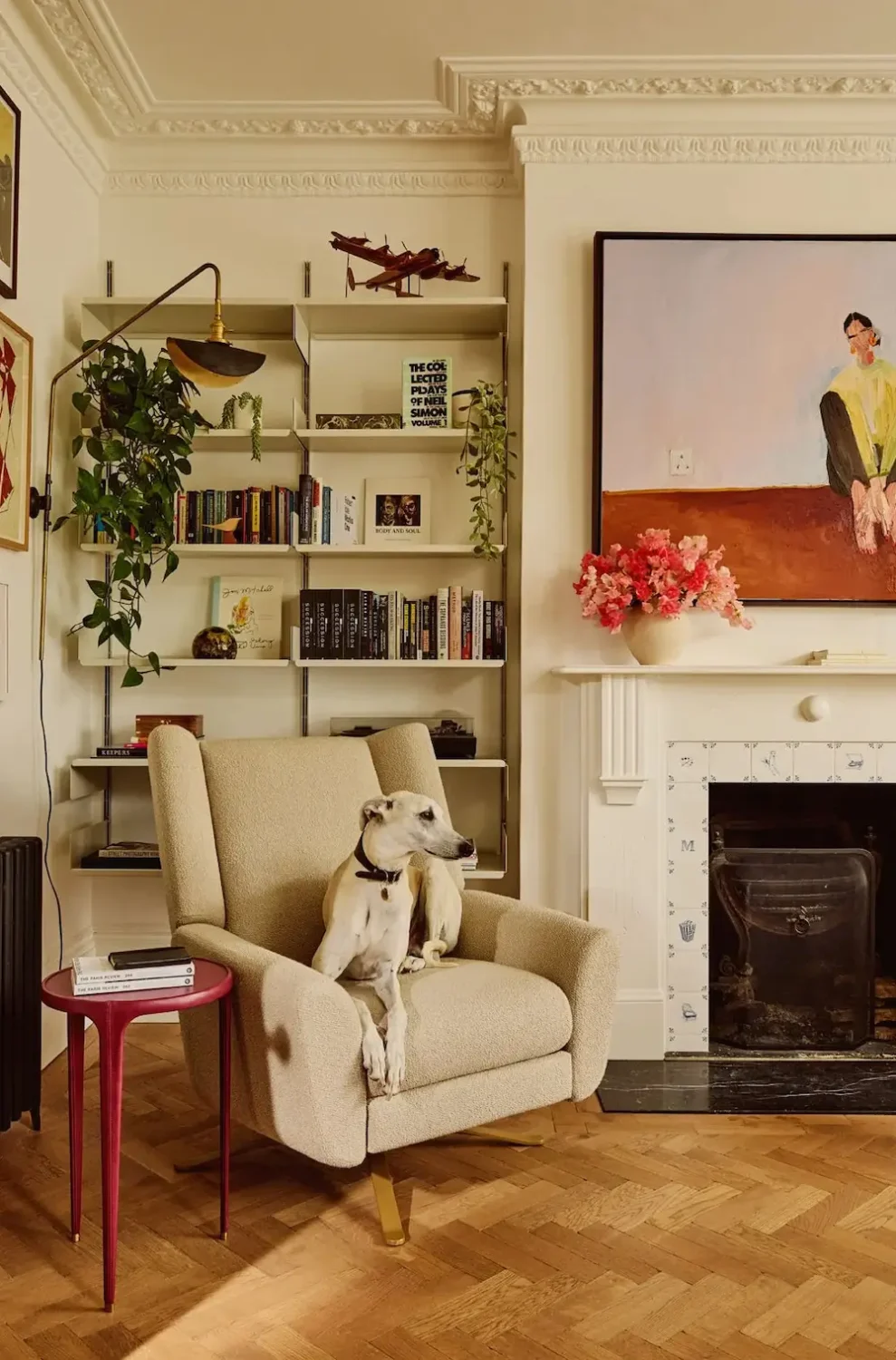 Reading nook in Michelle Dockery's London Victorian home featuring a cream bouclé armchair, open bookshelves, a white fireplace with decorative tiles, herringbone wood flooring, and a greyhound resting in the chair.
