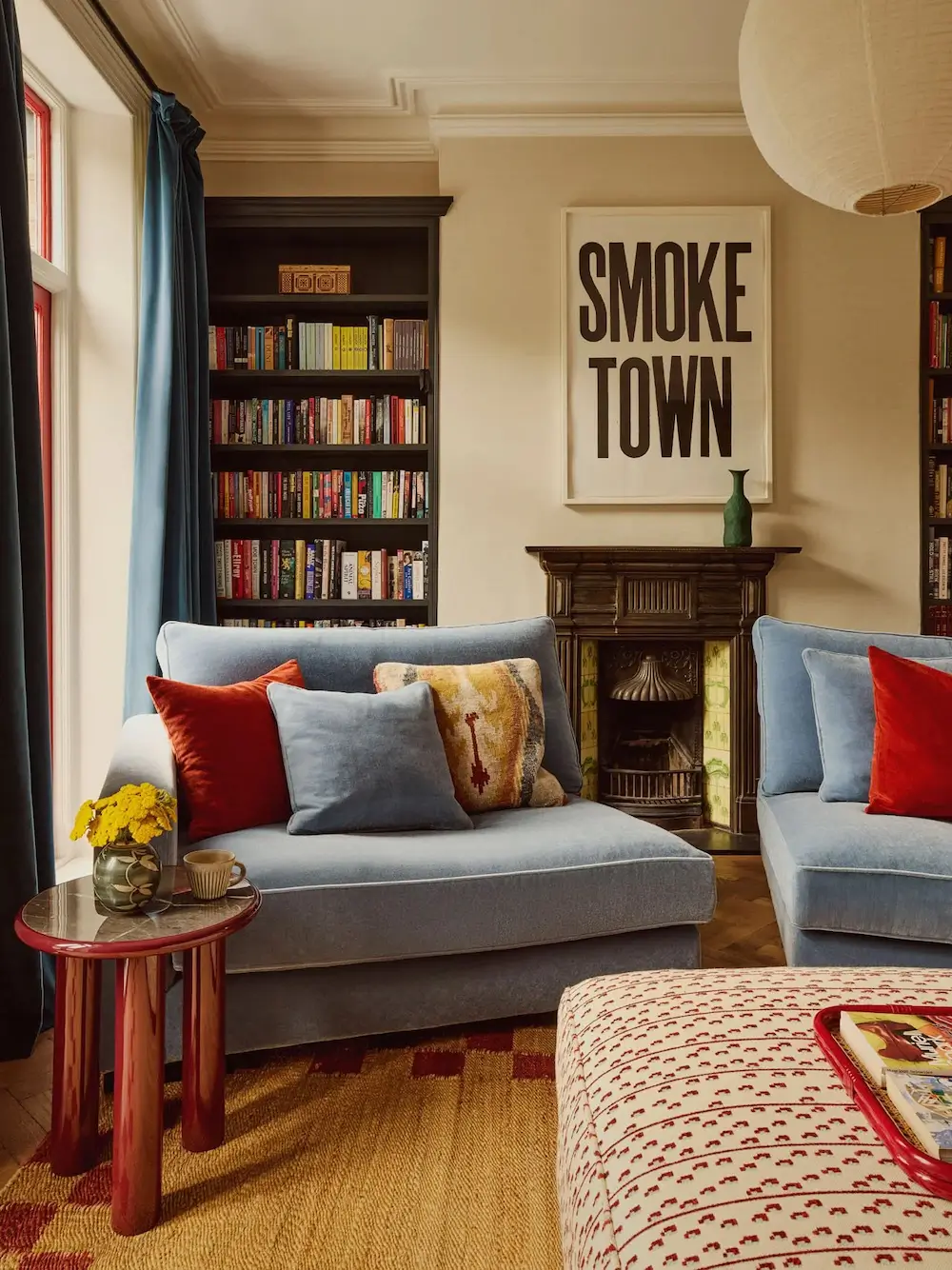 Sitting room in Michelle Dockery's London Victorian home featuring pale blue sofas, a vintage tiled fireplace, dark built-in bookshelves, a red side table, and bold typography artwork.