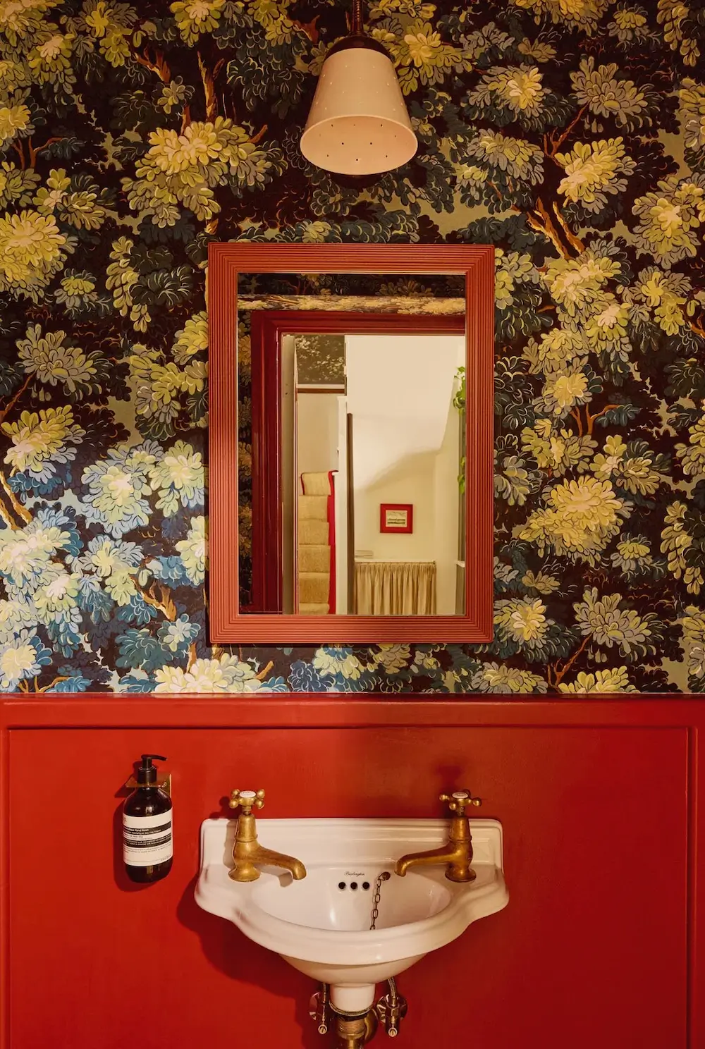 Powder room in Michelle Dockery's London Victorian home featuring rich red wainscoting, woodland wallpaper, a vintage wall-mounted sink with brass taps, and a red-framed mirror designed by Emma Ainscough.