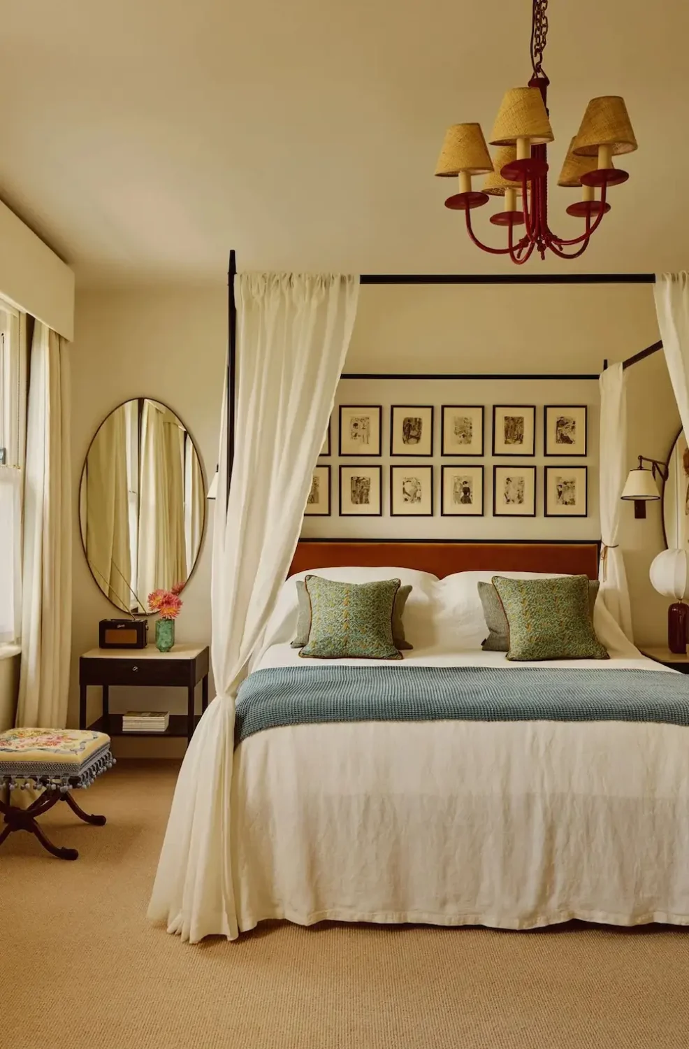Primary bedroom in Michelle Dockery's London Victorian home featuring a black four-poster bed with white linen curtains, blue accent bedding, a gallery wall of framed artwork, and a red chandelier.