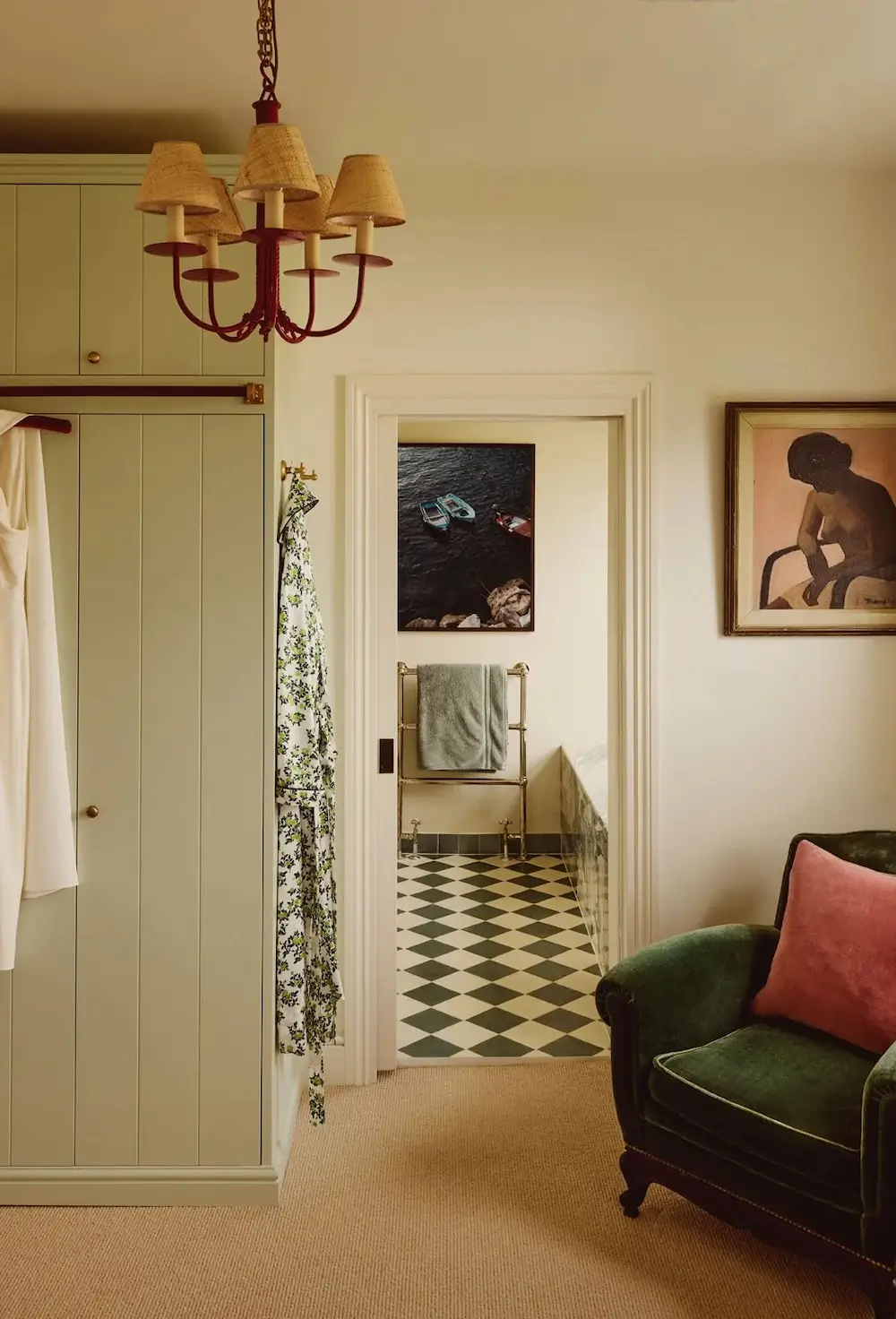Primary bedroom in Michelle Dockery's London Victorian home featuring sage green built-in wardrobes, a green velvet armchair, a red chandelier, and an en suite bathroom with a black-and-white checkerboard floor.