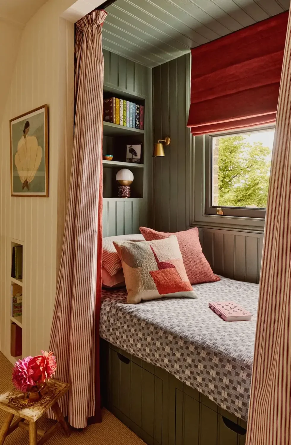 Built-in window seat in Michelle Dockery's London Victorian home featuring sage green paneling, striped curtains, a deep red Roman shade, open shelving, and pink accent cushions.