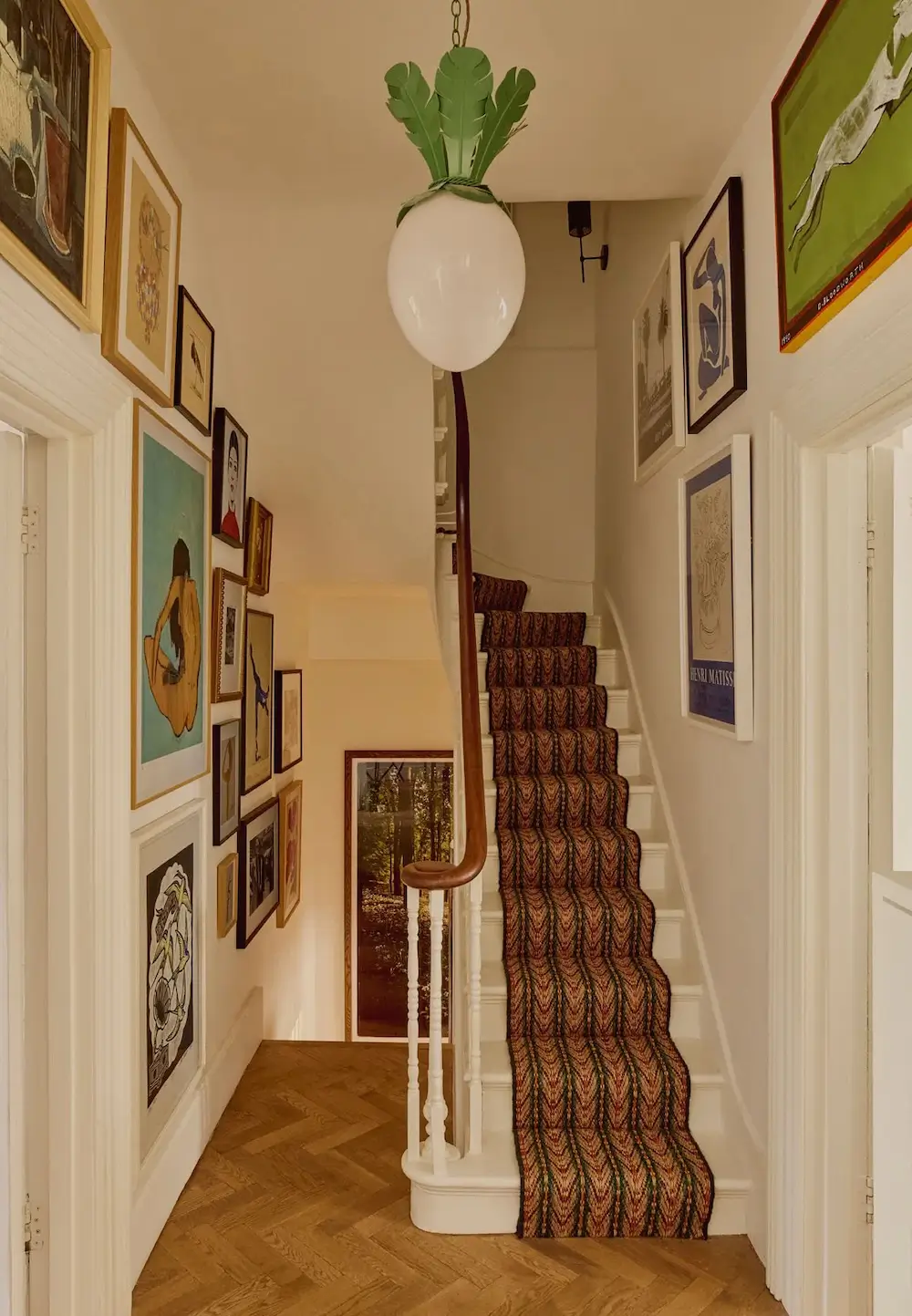 Hallway in Michelle Dockery's London Victorian home featuring a gallery wall of eclectic artwork, a patterned stair runner, herringbone wood flooring, and a whimsical radish-shaped pendant light.