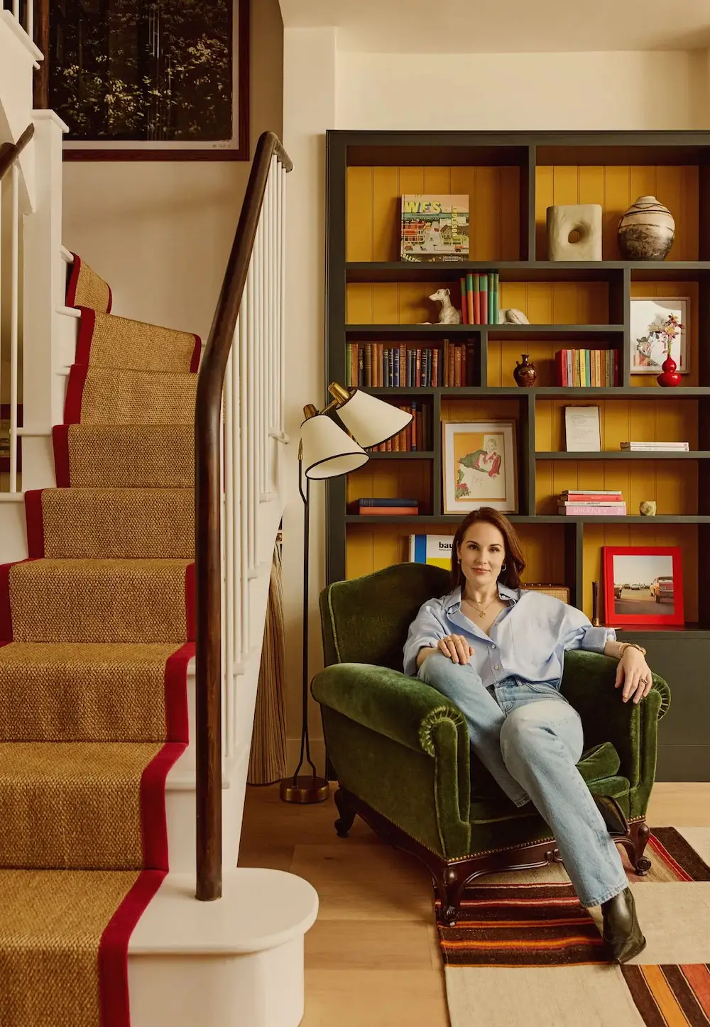 Michelle Dockery seated in a green velvet armchair beside a staircase with a red-edged runner, in front of bespoke shelving filled with books, artwork, and ceramics in her London Victorian home designed by Emma Ainscough.
