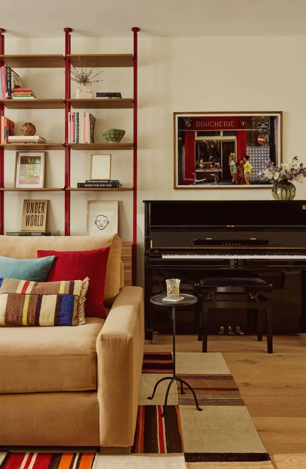 Living room in Michelle Dockery's London Victorian home featuring a beige sofa with colorful cushions, open shelving with red supports, a black upright piano, striped rug, and framed artwork.