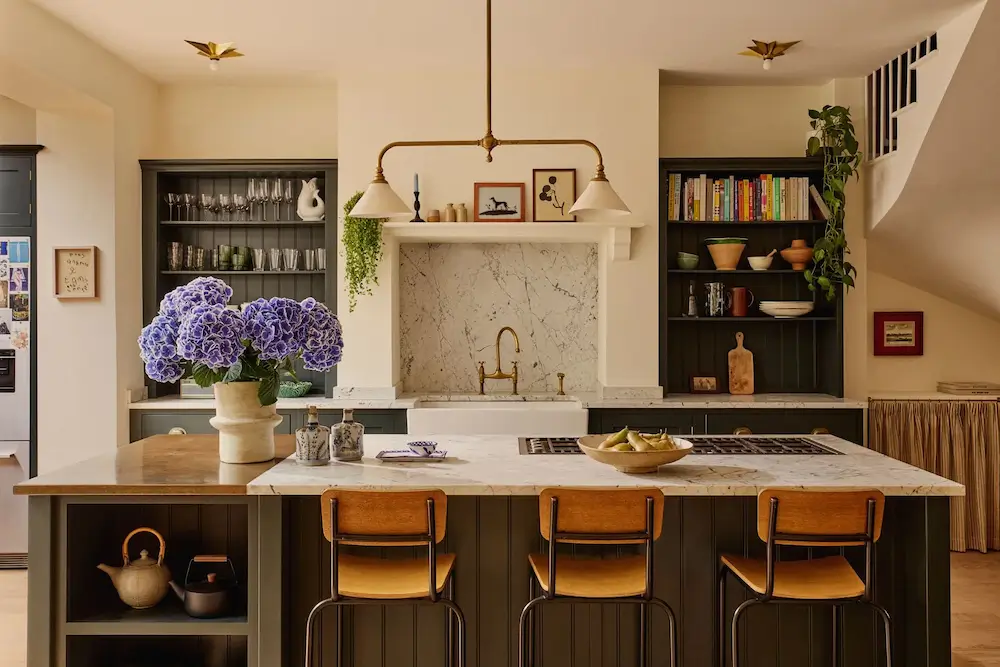 Kitchen in Michelle Dockery's London Victorian home featuring a marble island with wooden bar stools, dark green cabinetry, open shelving, a farmhouse sink, and brass lighting designed by Emma Ainscough.