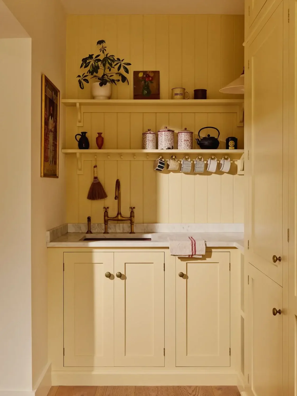 Butler's pantry in Michelle Dockery's London Victorian home with cream shaker cabinetry, marble countertop, brass faucet, open shelves displaying mugs and vintage canisters, and tongue-and-groove paneling.