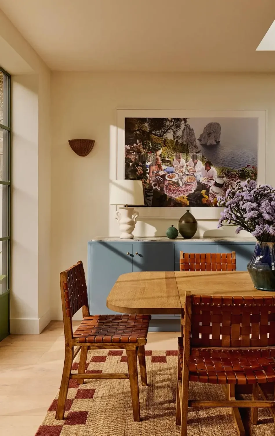 Dining room in Michelle Dockery's London Victorian home featuring a solid oak dining table, woven leather chairs, a blue sideboard, oversized coastal artwork, and fresh lilac flowers.