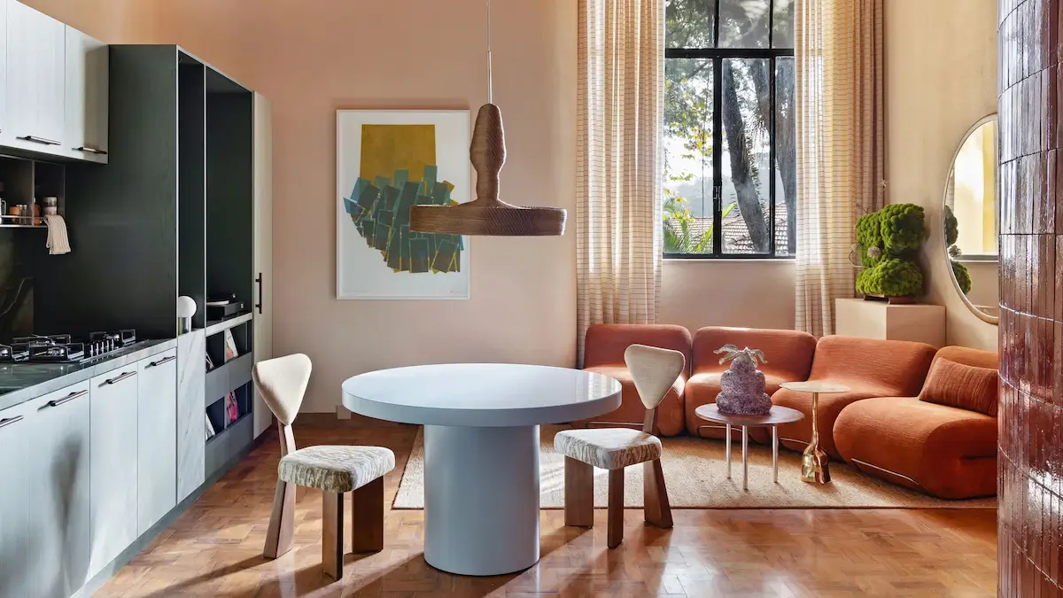 Open-plan living room and kitchen in a small Brazilian apartment featuring warm terracotta sofas, a round white dining table, parquet flooring, floor-to-ceiling windows, sculptural lighting, and natural textures.