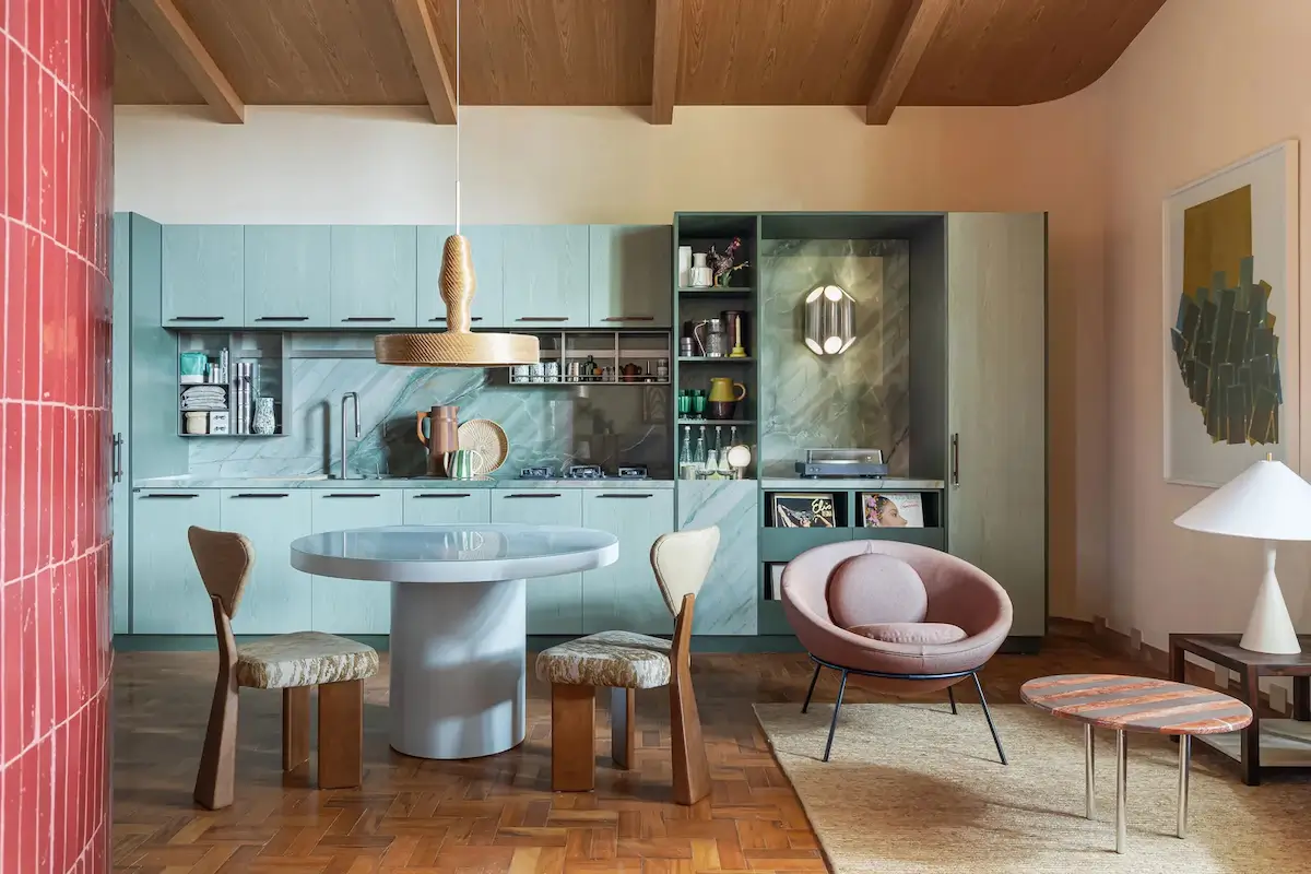Modern kitchen and dining area in a small Brazilian apartment with sage green cabinetry, a marble backsplash, a round pedestal dining table, exposed timber ceiling beams, and a blush accent chair.